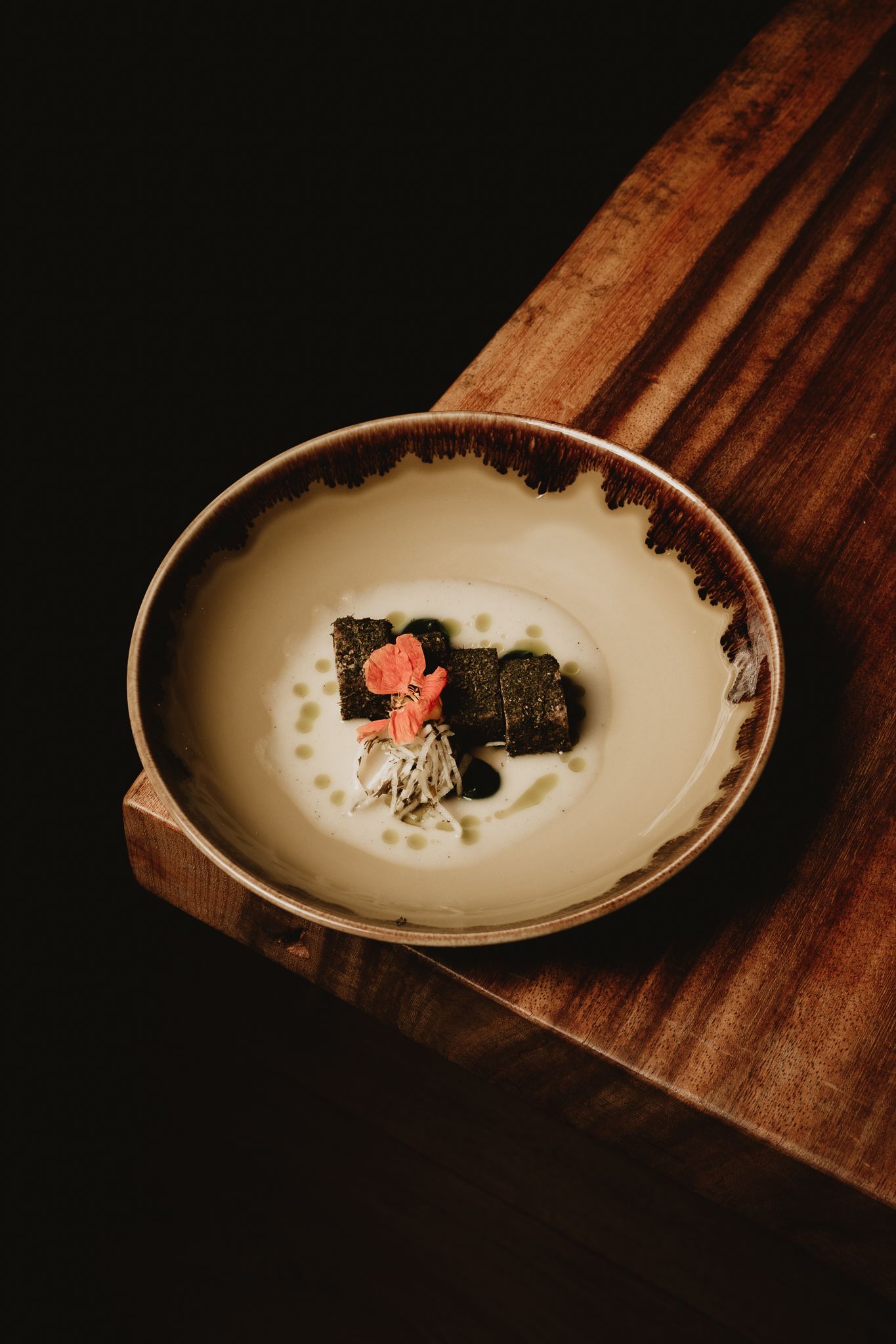 Elegant plated dish with nori-wrapped filling, pink flower garnish, and dots of sauce on a ceramic plate atop a wooden surface.