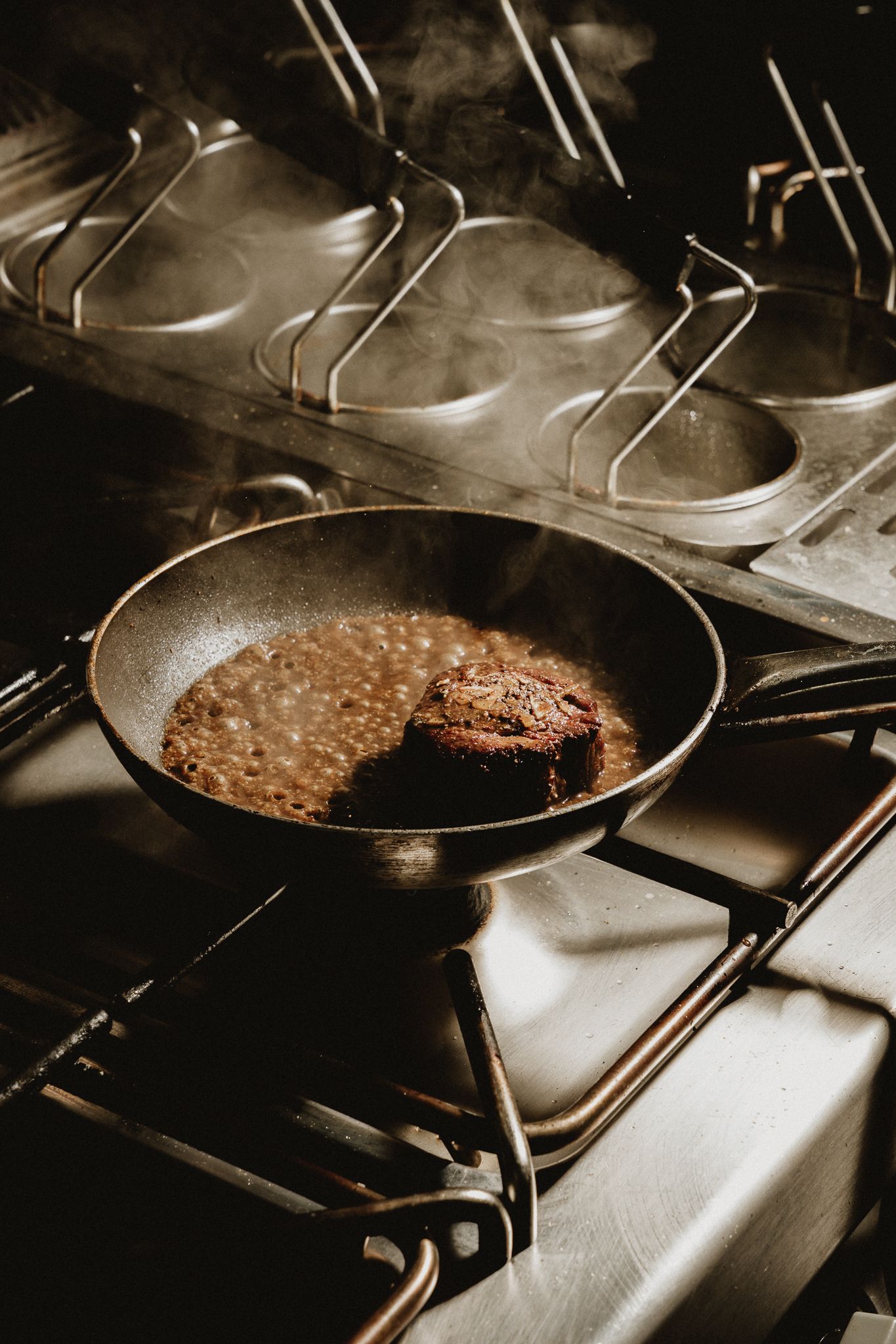 A juicy beef patty sizzling in a cast iron skillet on a commercial stainless steel stove with steam rising.