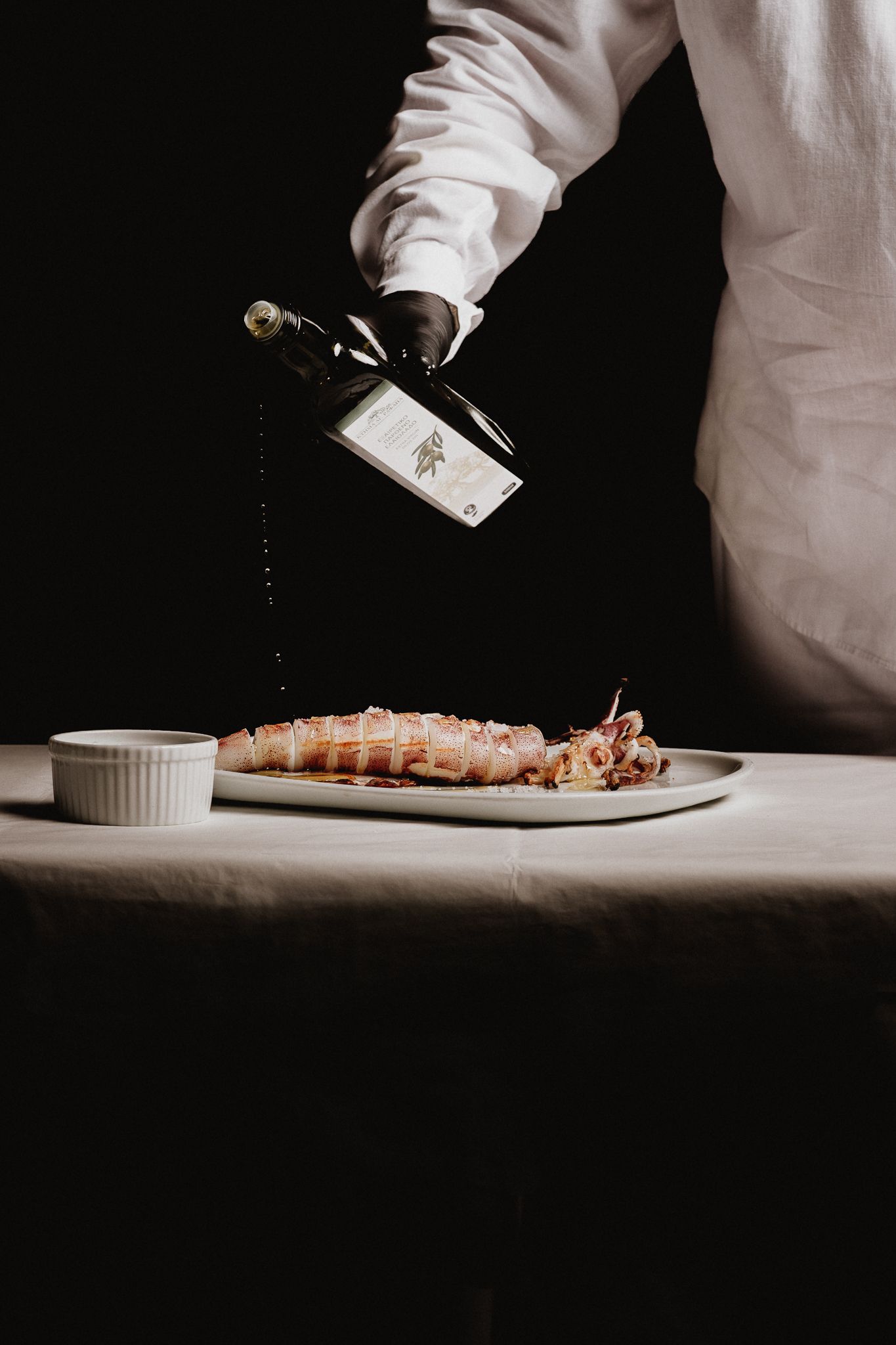Chef drizzles olive oil over an elegantly plated meat dish on a white tablecloth.