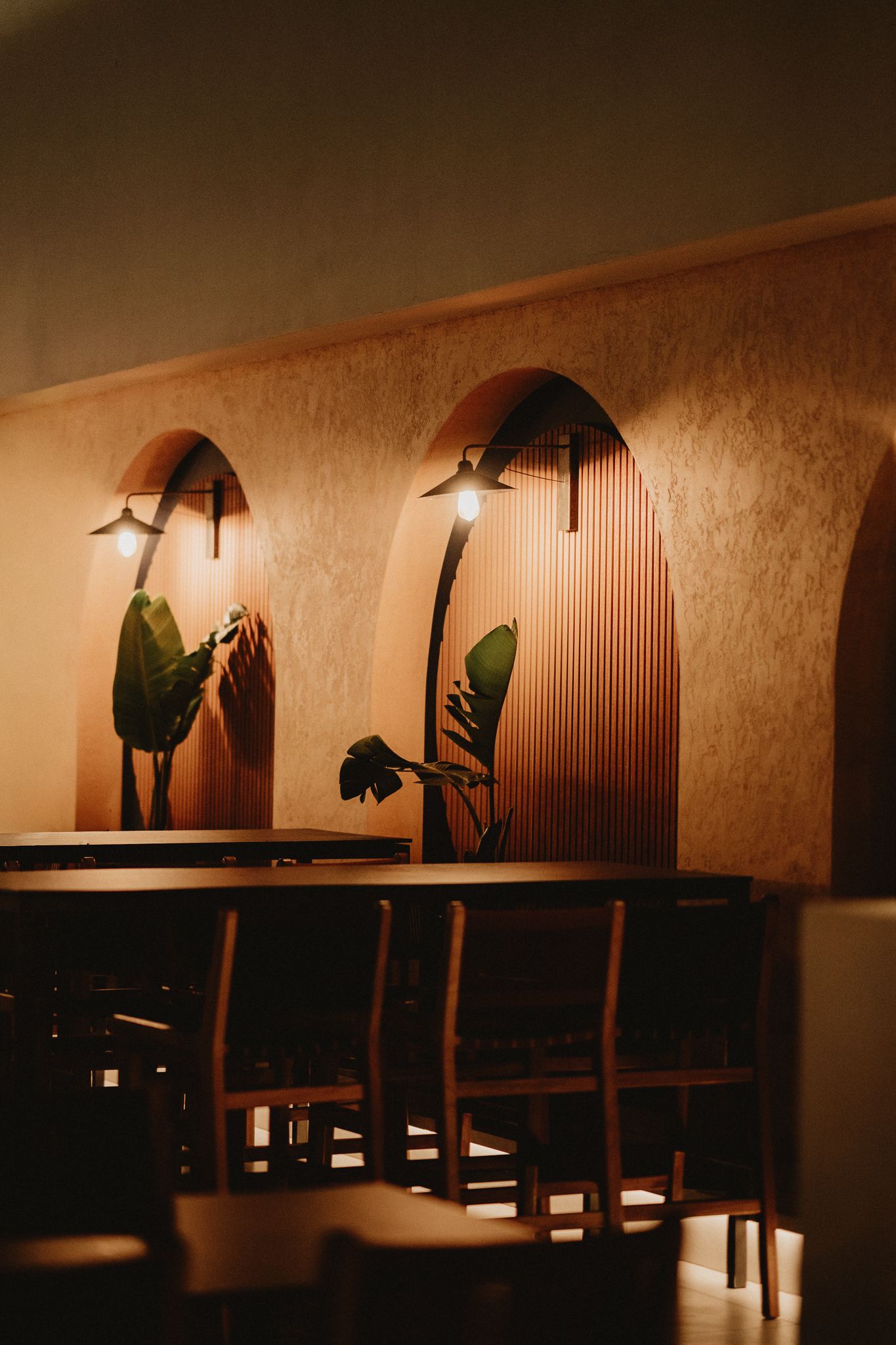 Warm restaurant interior with arched alcoves, wall sconces, tropical plants, and wooden bar seating in moody amber lighting.