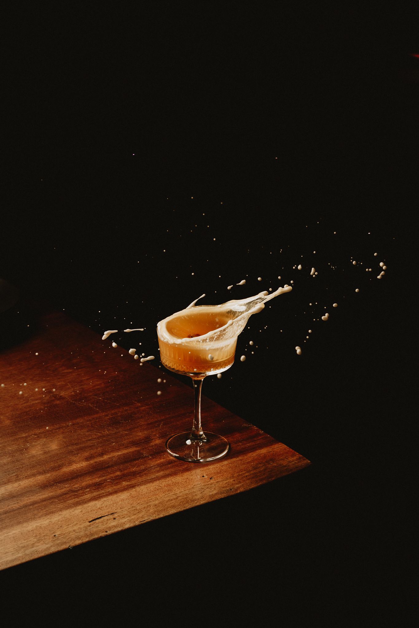 A cocktail splashing dramatically in an elegant coupe glass on a wooden surface against a dark background.