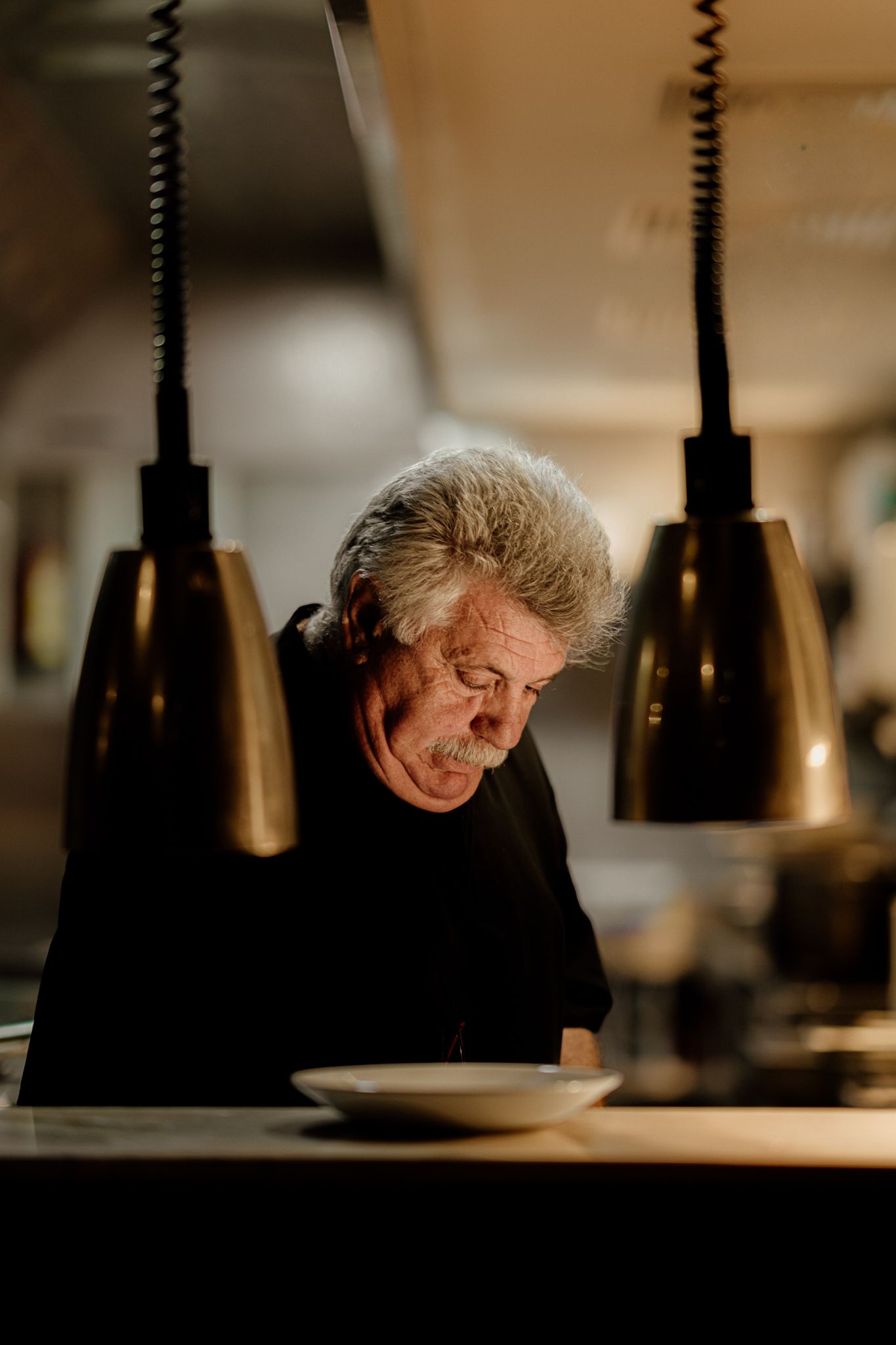Elderly chef with gray hair working under warm hanging lamps in a dimly lit restaurant kitchen.