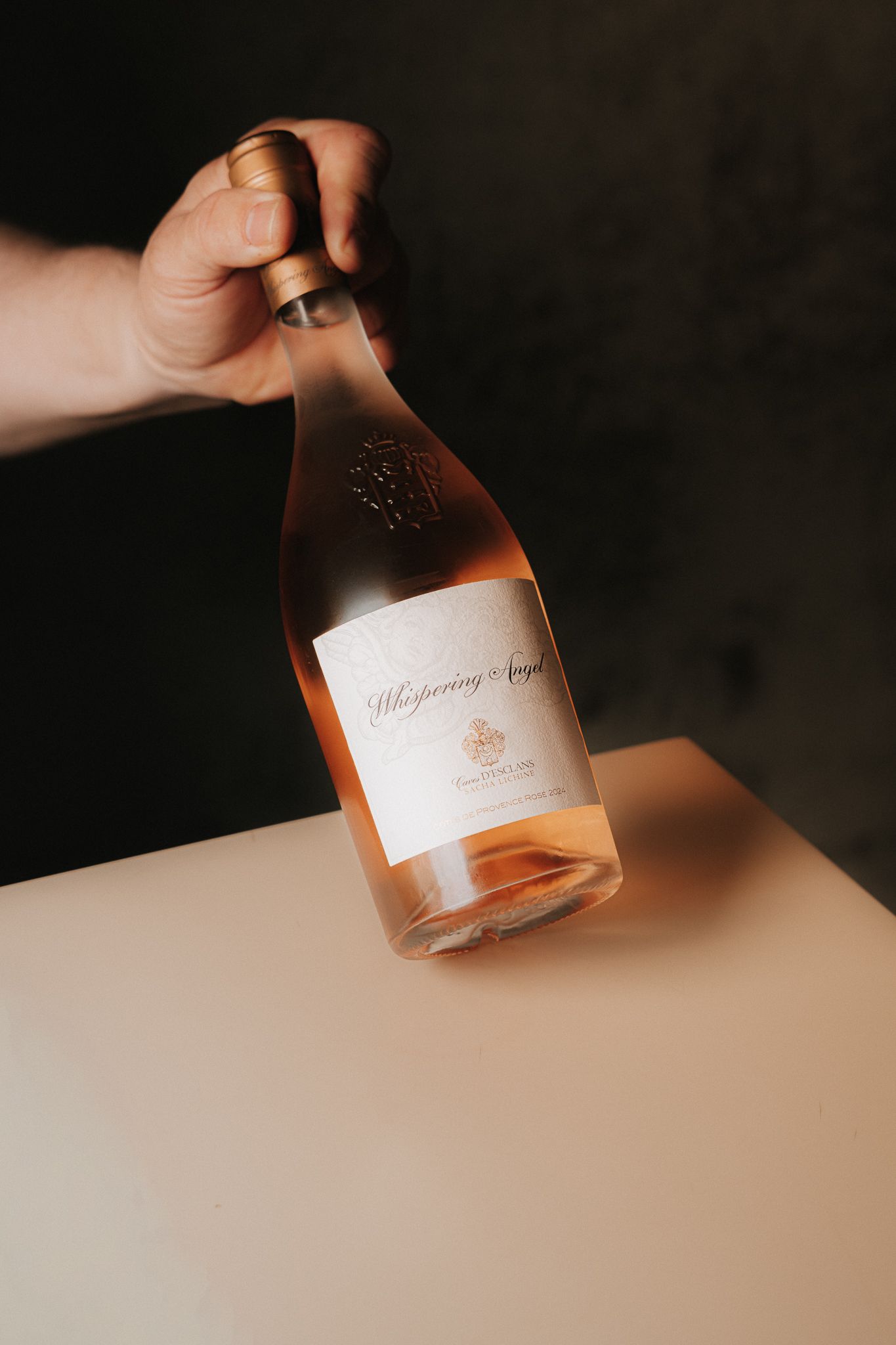 Hand holding a bottle of Whispering Angel rosé wine against a dark background.