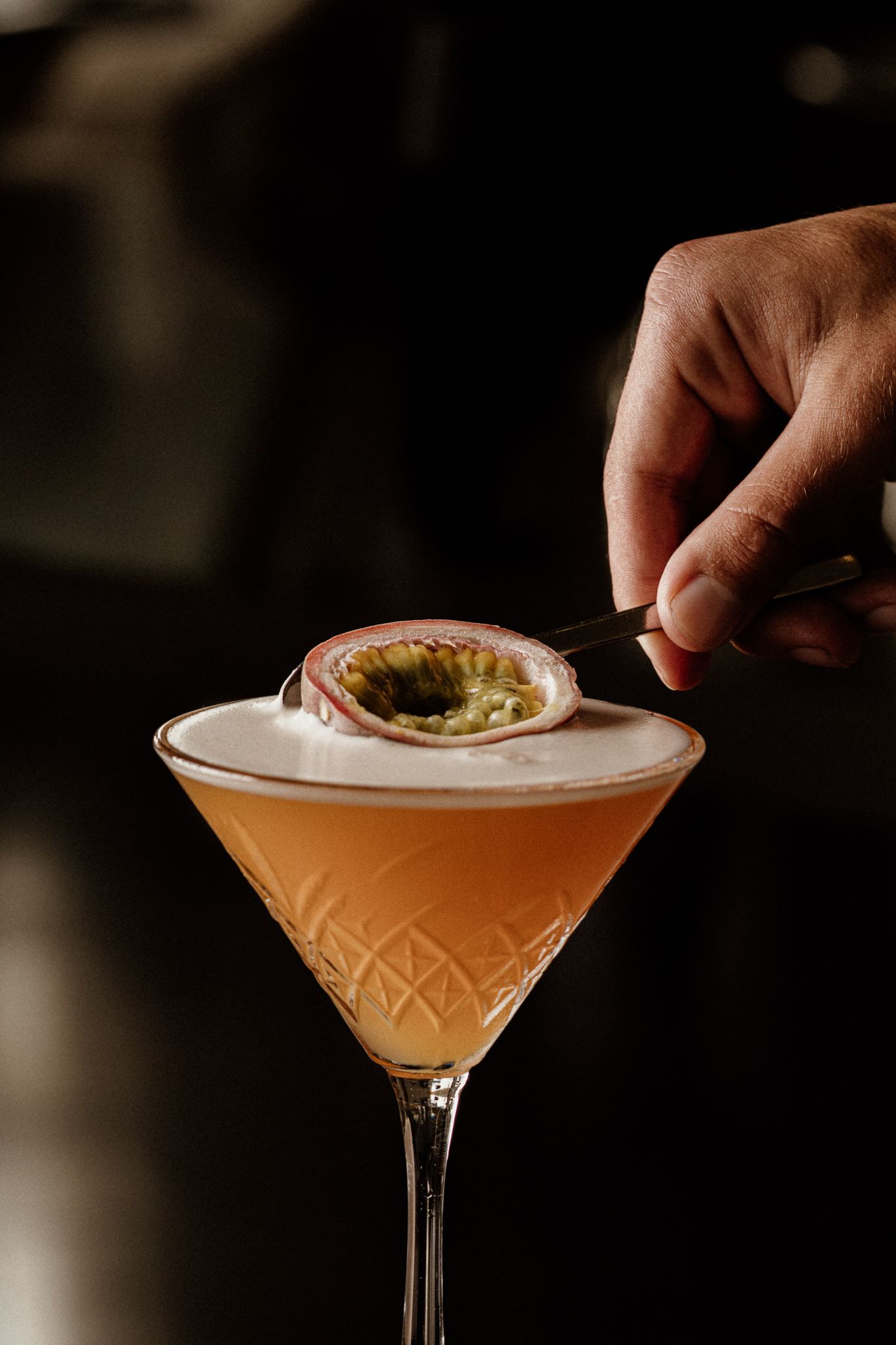 Bartender garnishing a frothy passion fruit martini cocktail in an elegant crystal glass on dark background.
