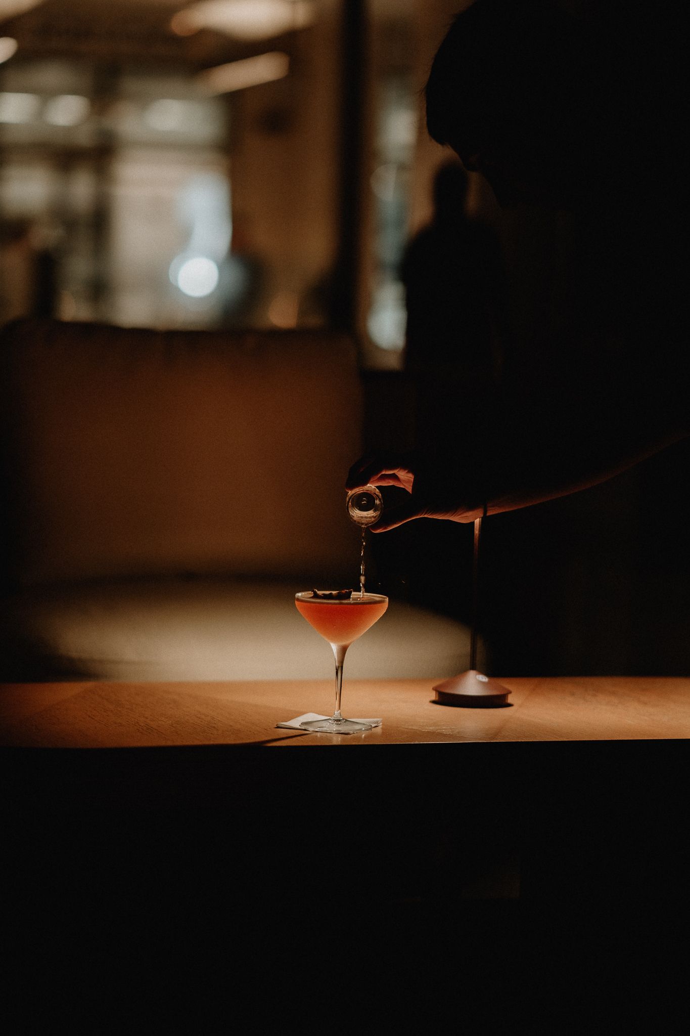 Bartender pouring a cocktail into an elegant coupe glass on a dimly lit bar counter.