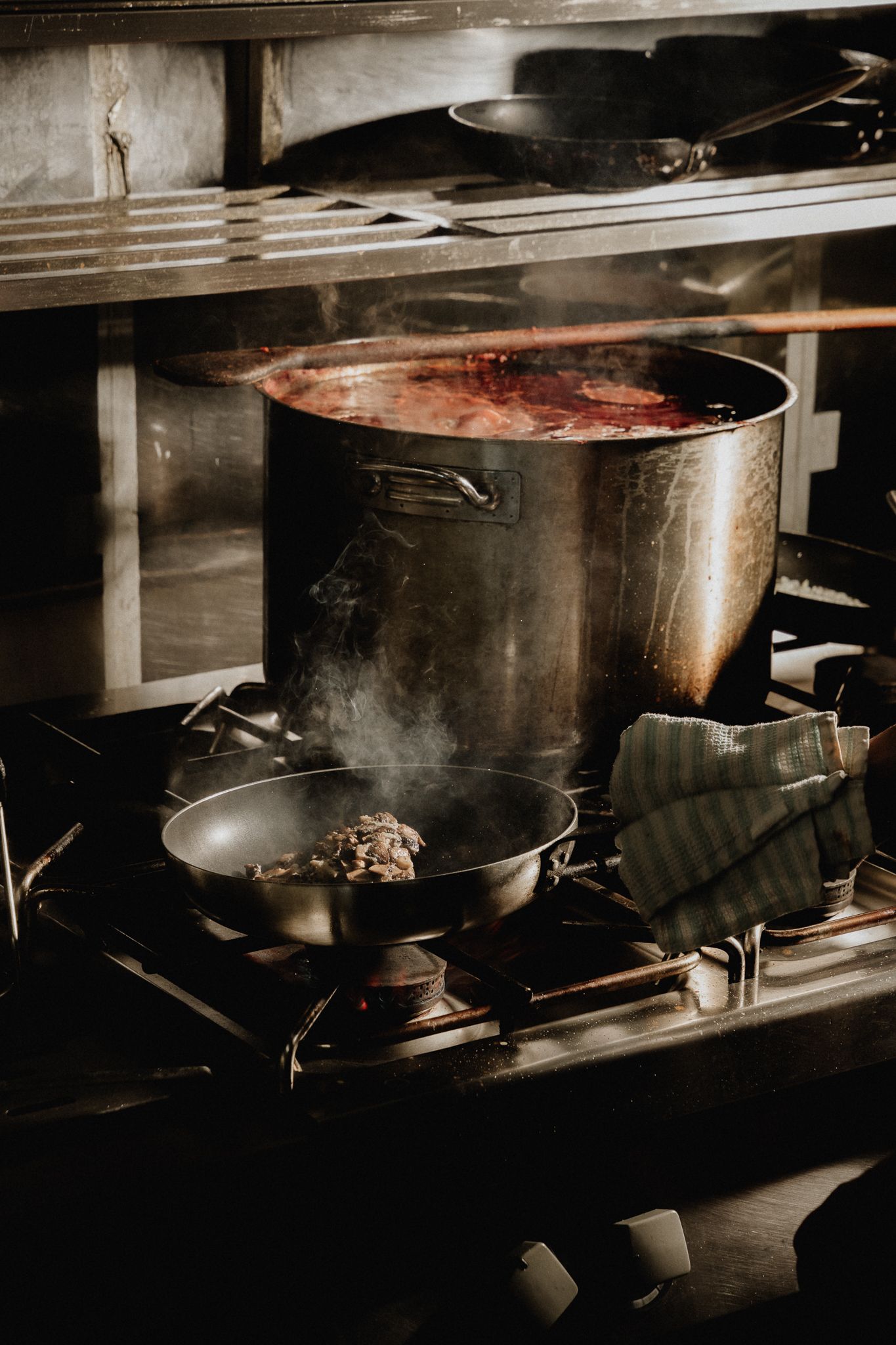 Large stockpot with red sauce simmering on commercial gas stove alongside small frying pan in dark kitchen.