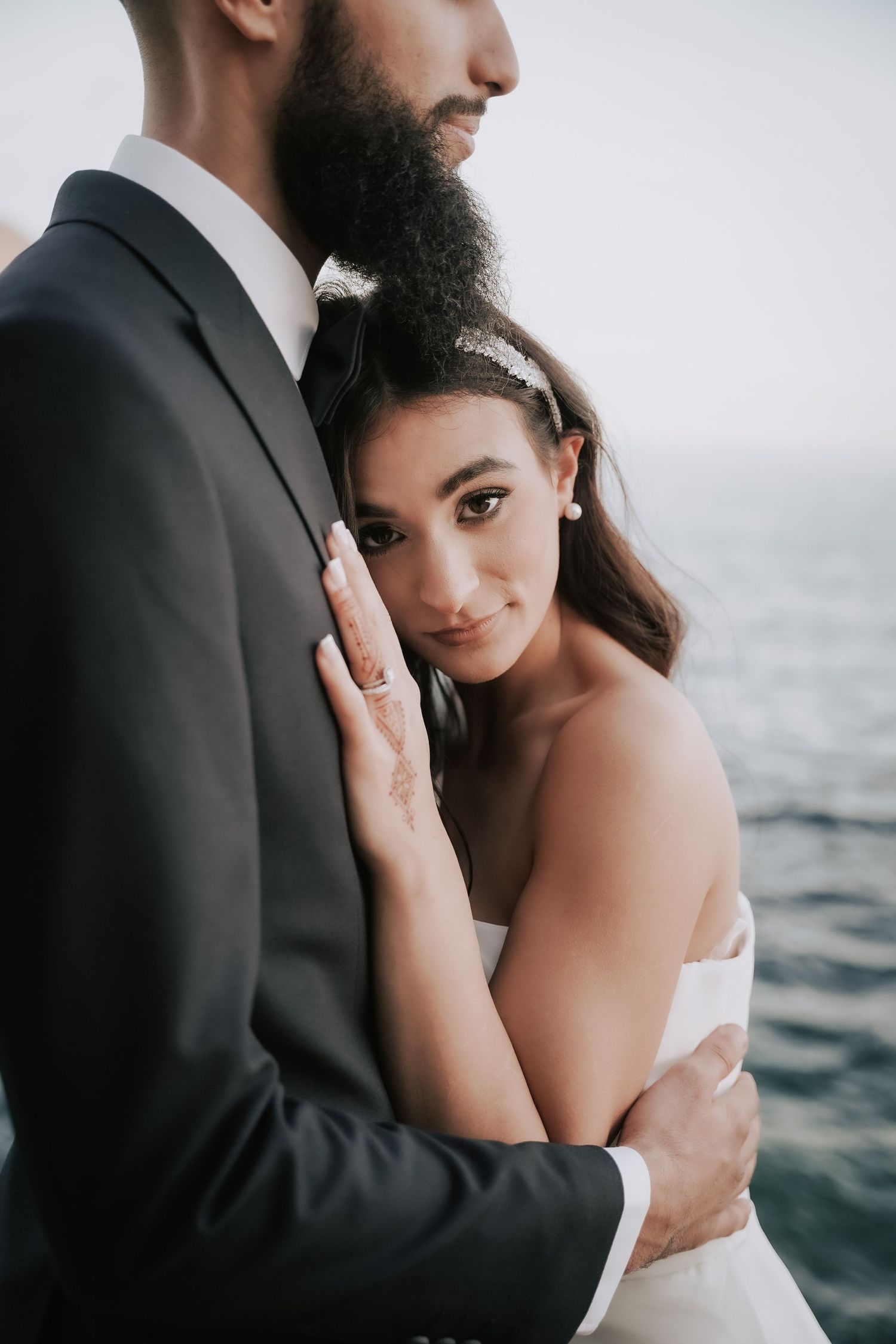 Bride with henna tattoos embraces bearded groom in black suit beside a stunning ocean backdrop.