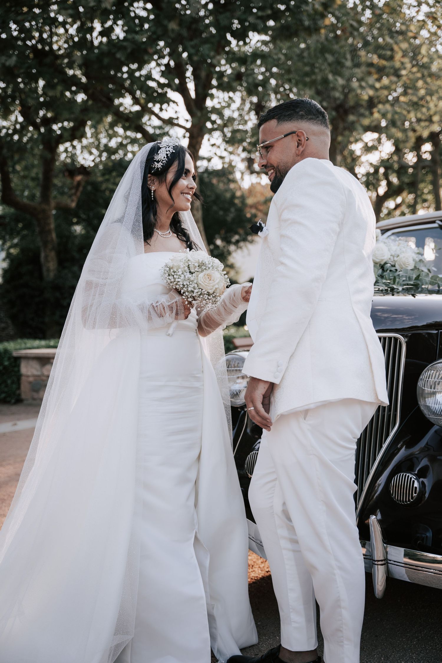 Bride in white gown and groom in ivory suit smile together beside a classic vintage black car.