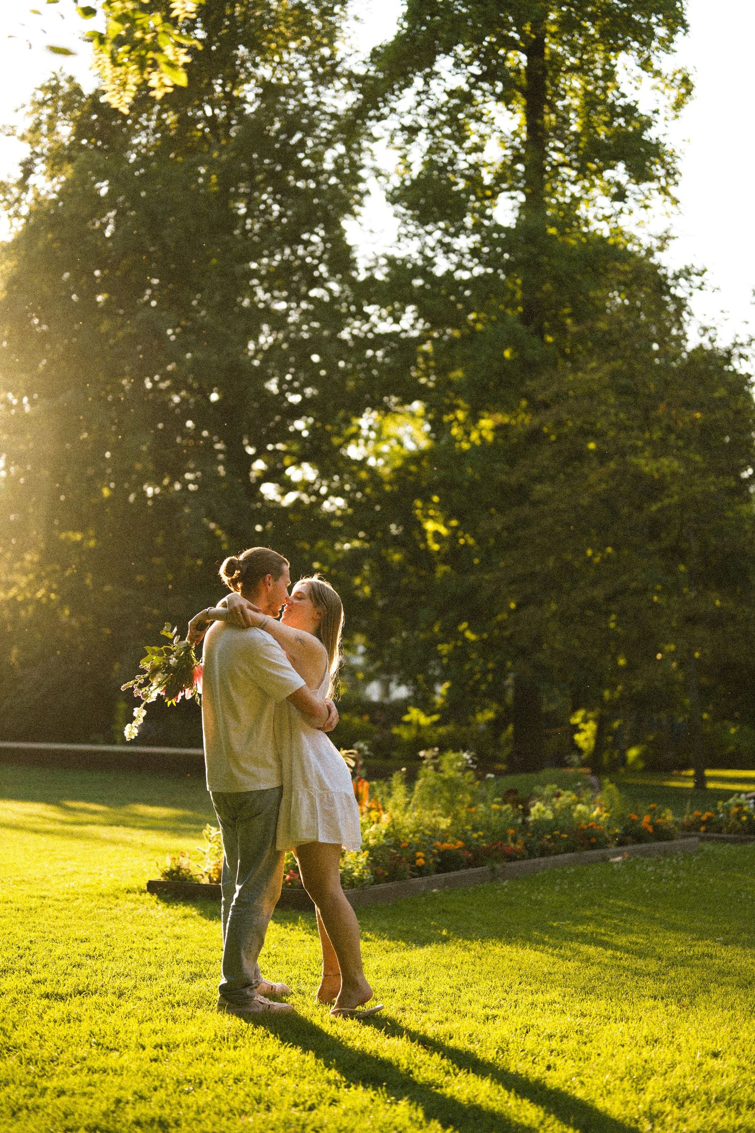 Romantic couple embracing and kissing in a golden sunlit park with tall green trees in the background.