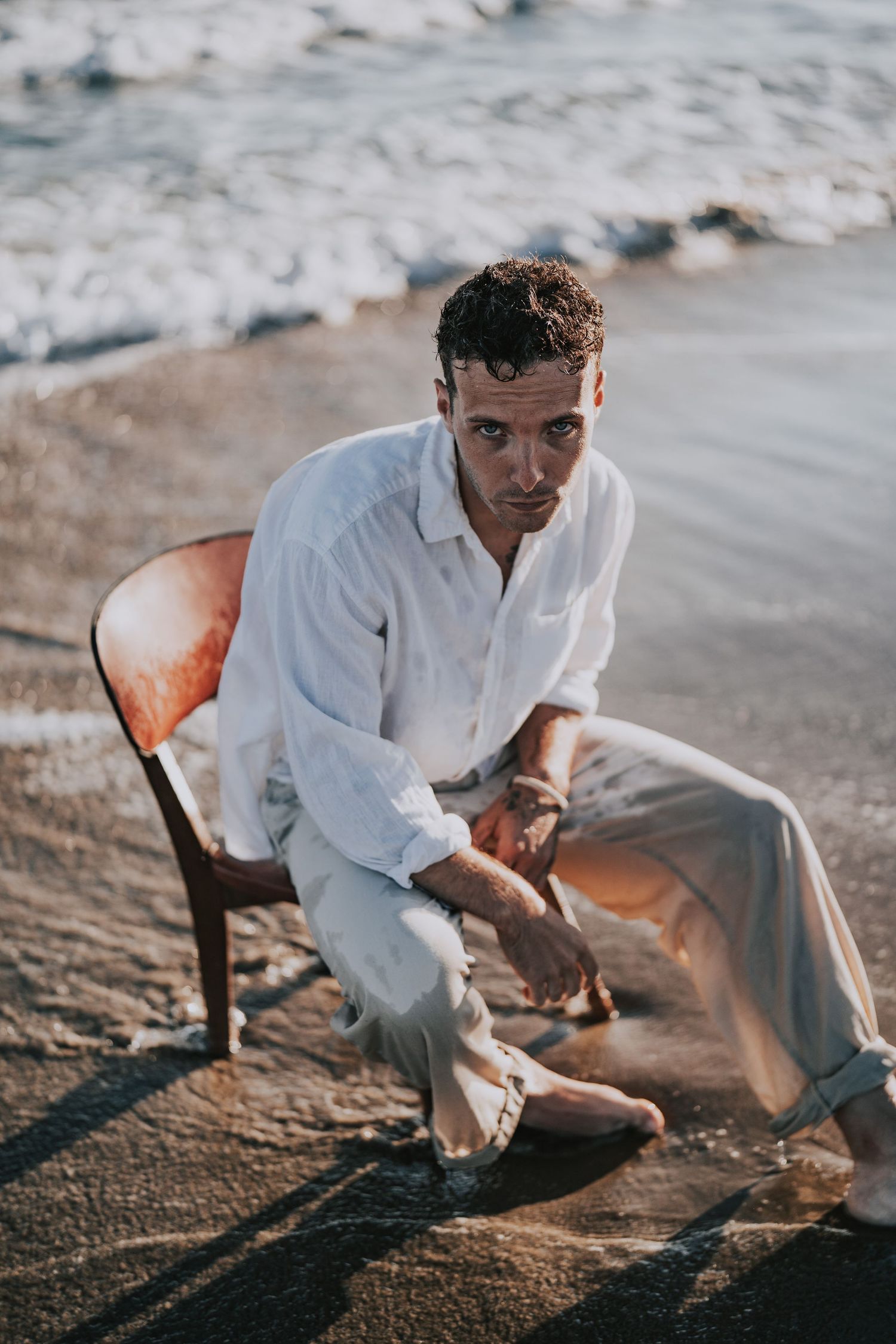 Man in white linen shirt sitting on chair at beach shoreline with ocean waves behind him.