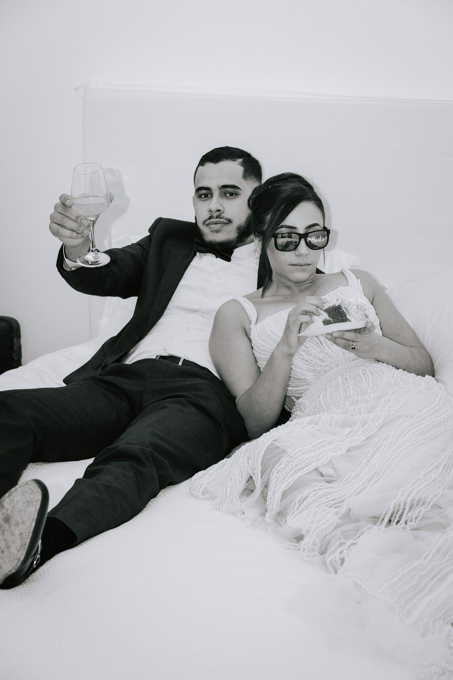 Black and white photo of newlyweds relaxing on bed, groom raising champagne glass, bride in sunglasses holding a photo.
