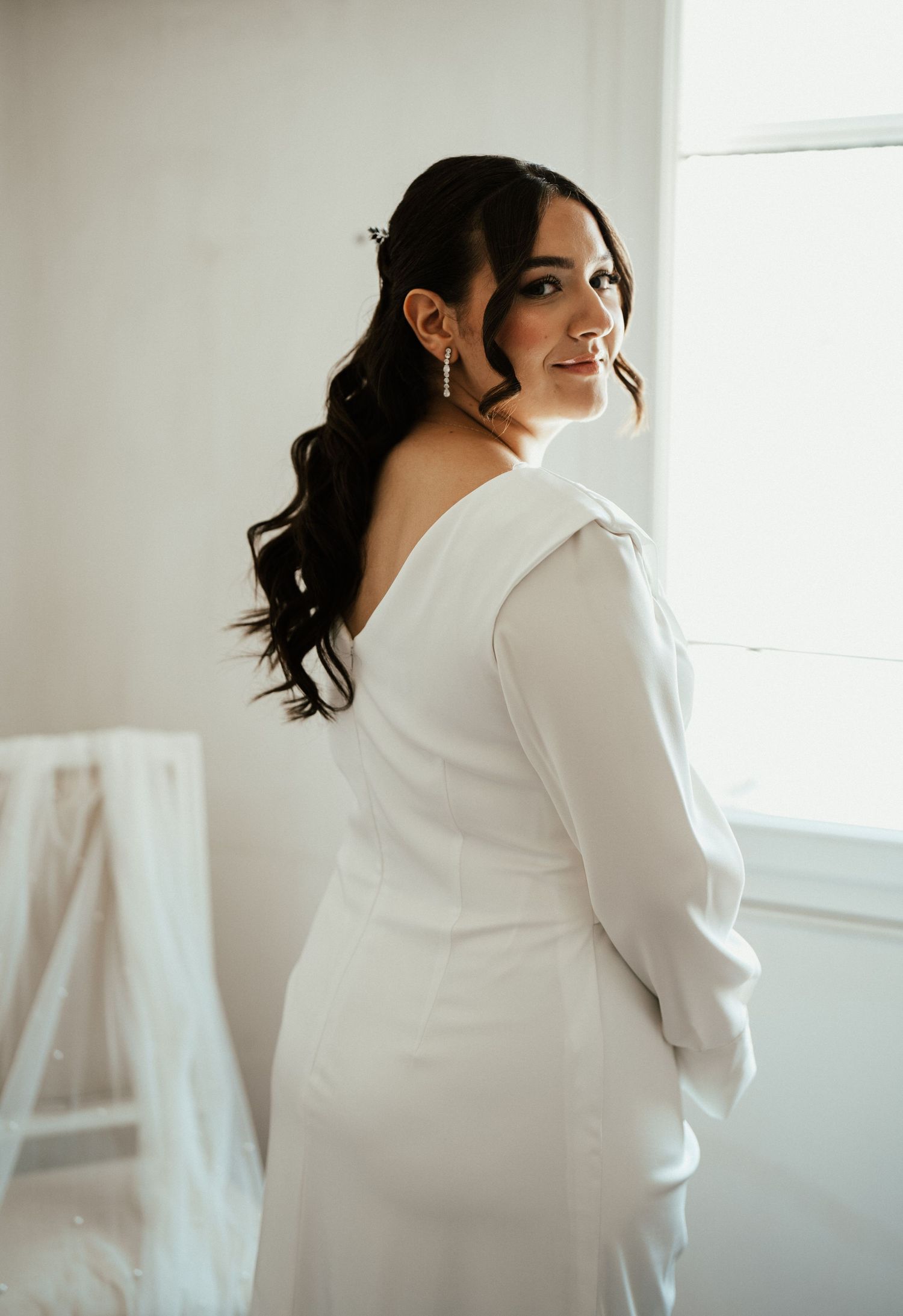 Bride in elegant white long-sleeve backless wedding gown posing gracefully near a bright window.