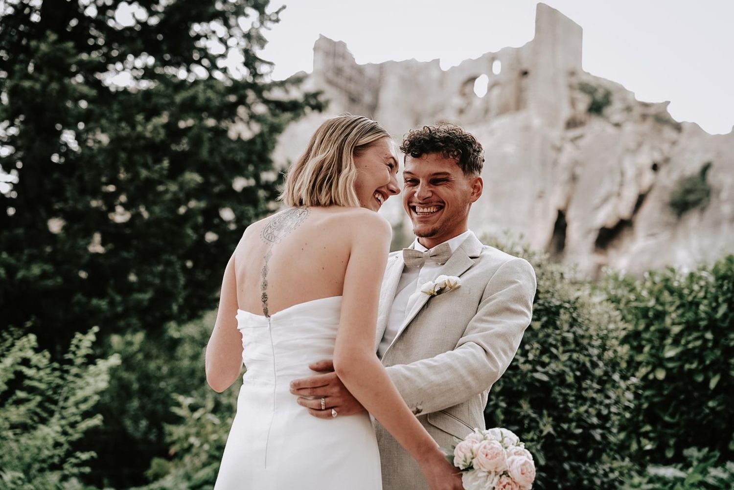 Laughing bride and groom embrace outdoors with ancient stone ruins behind them, he holding a pink bouquet.