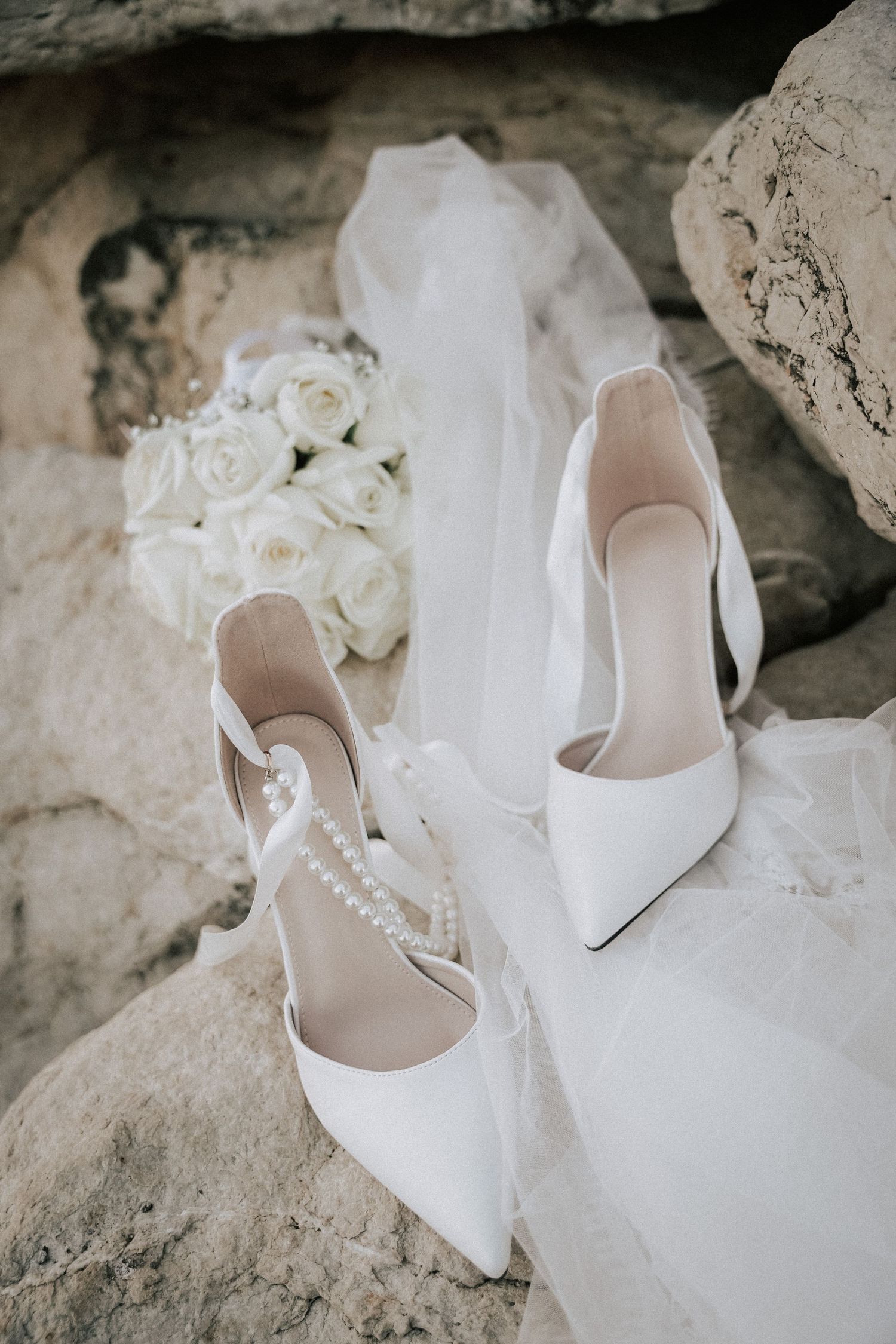 White bridal heels with pearl ankle straps beside a white rose bouquet and veil on rocky surface.
