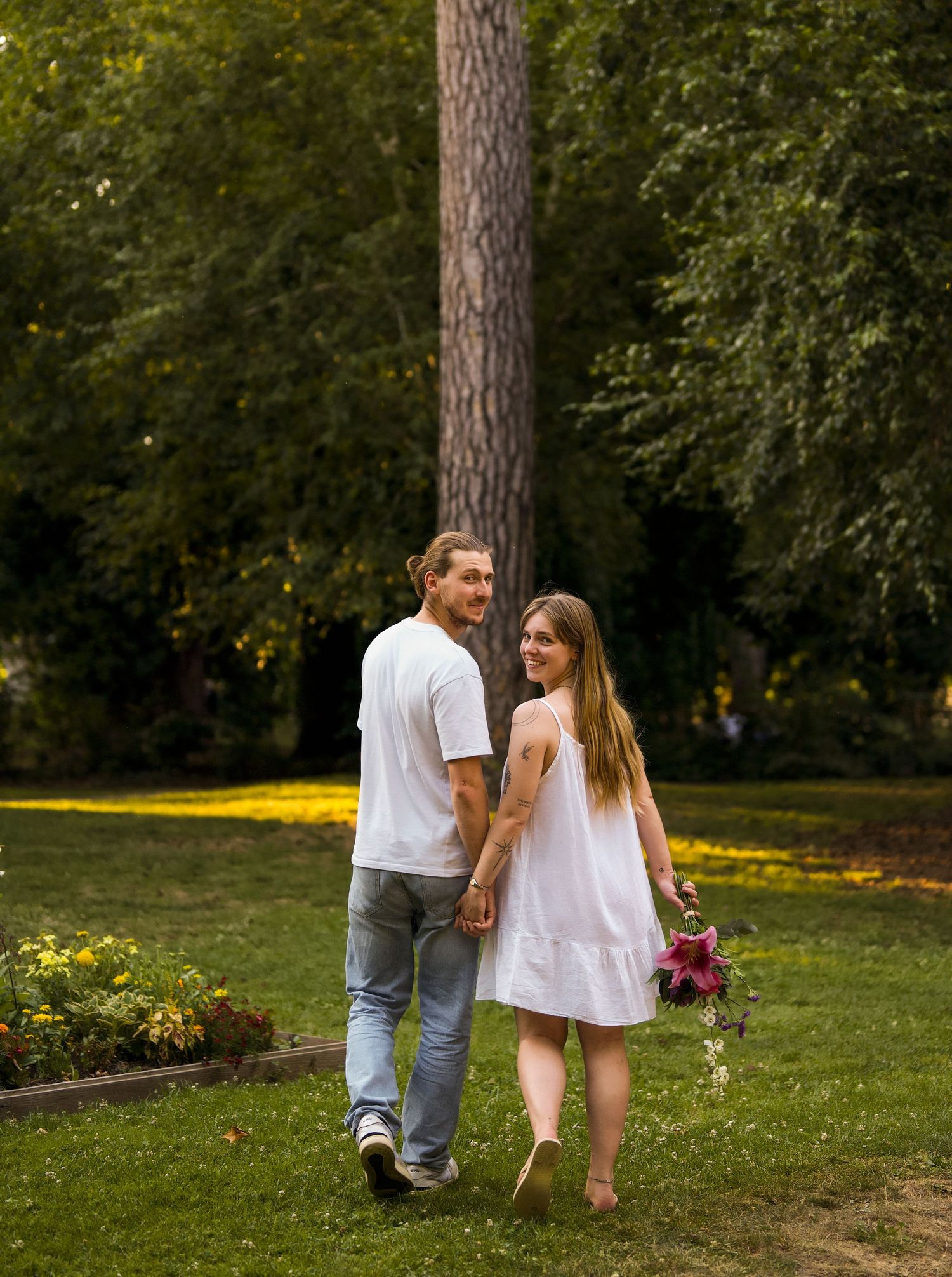 Couple walking hand in hand through a lush green park, woman carrying a bouquet of flowers.