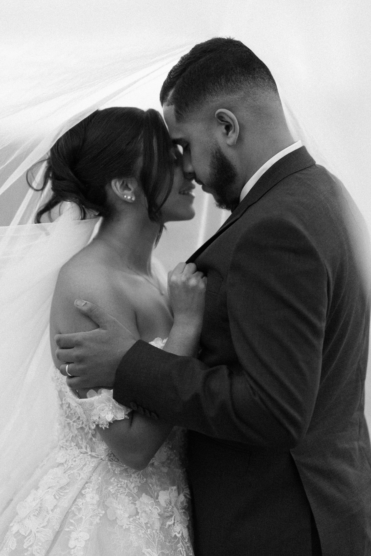Black and white romantic portrait of bride and groom touching foreheads under flowing veil in close embrace.