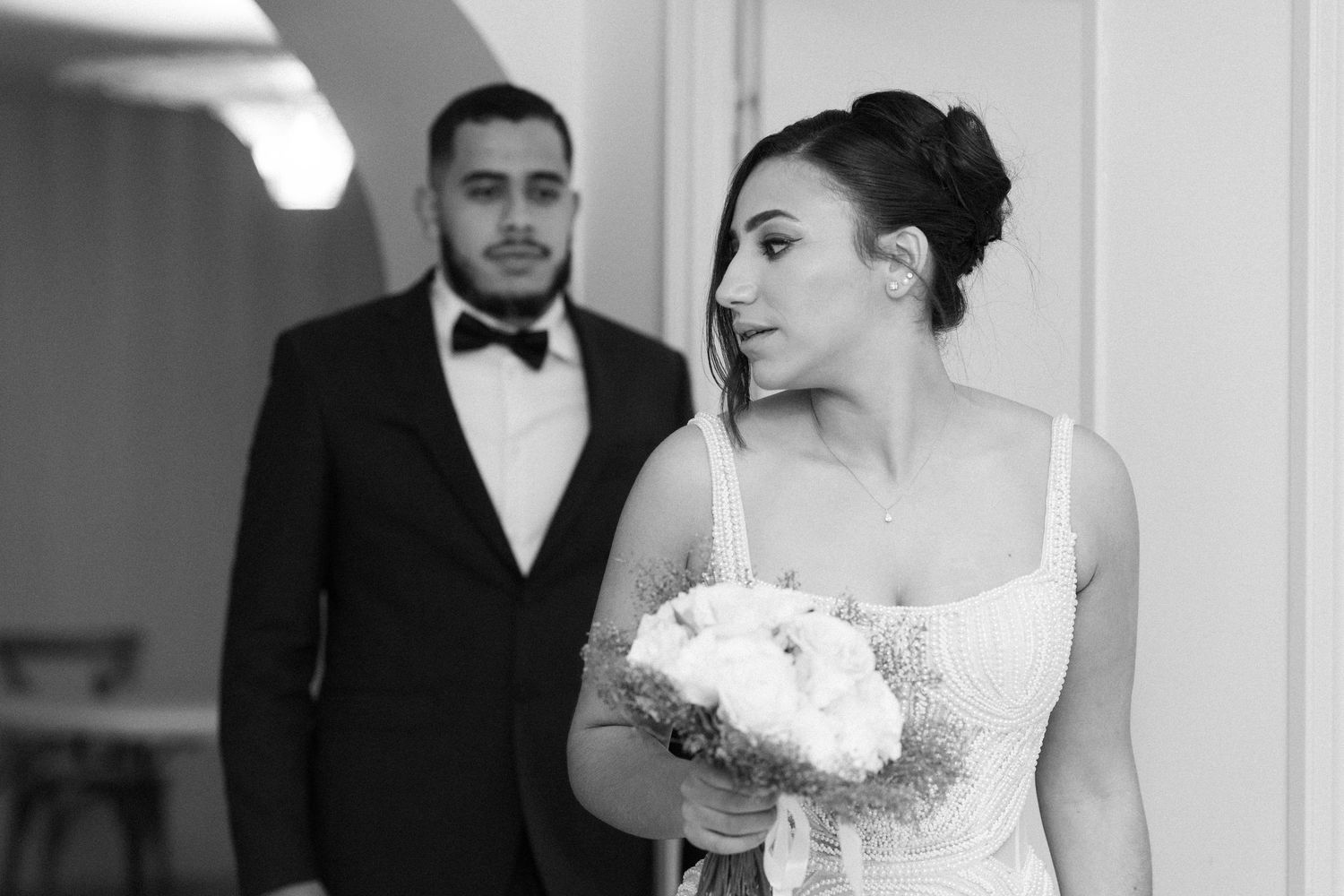 Black and white photo of bride holding bouquet walking ahead of groom in bow tie at wedding venue.