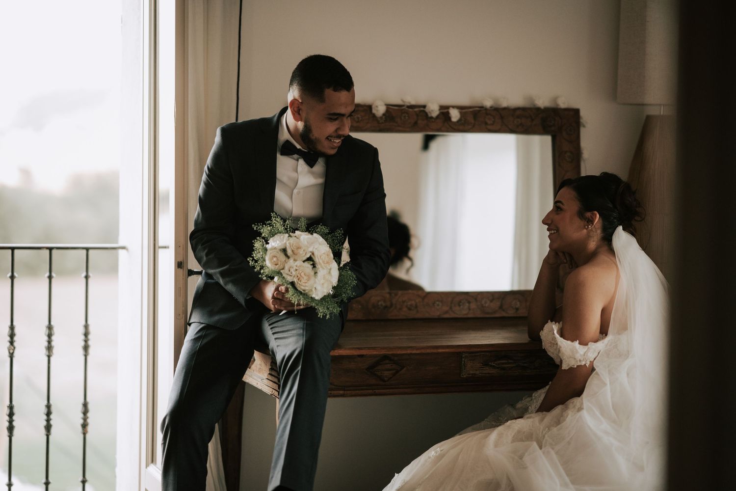 Groom in dark suit presents white bouquet to smiling bride in off-shoulder gown near vintage mirror.