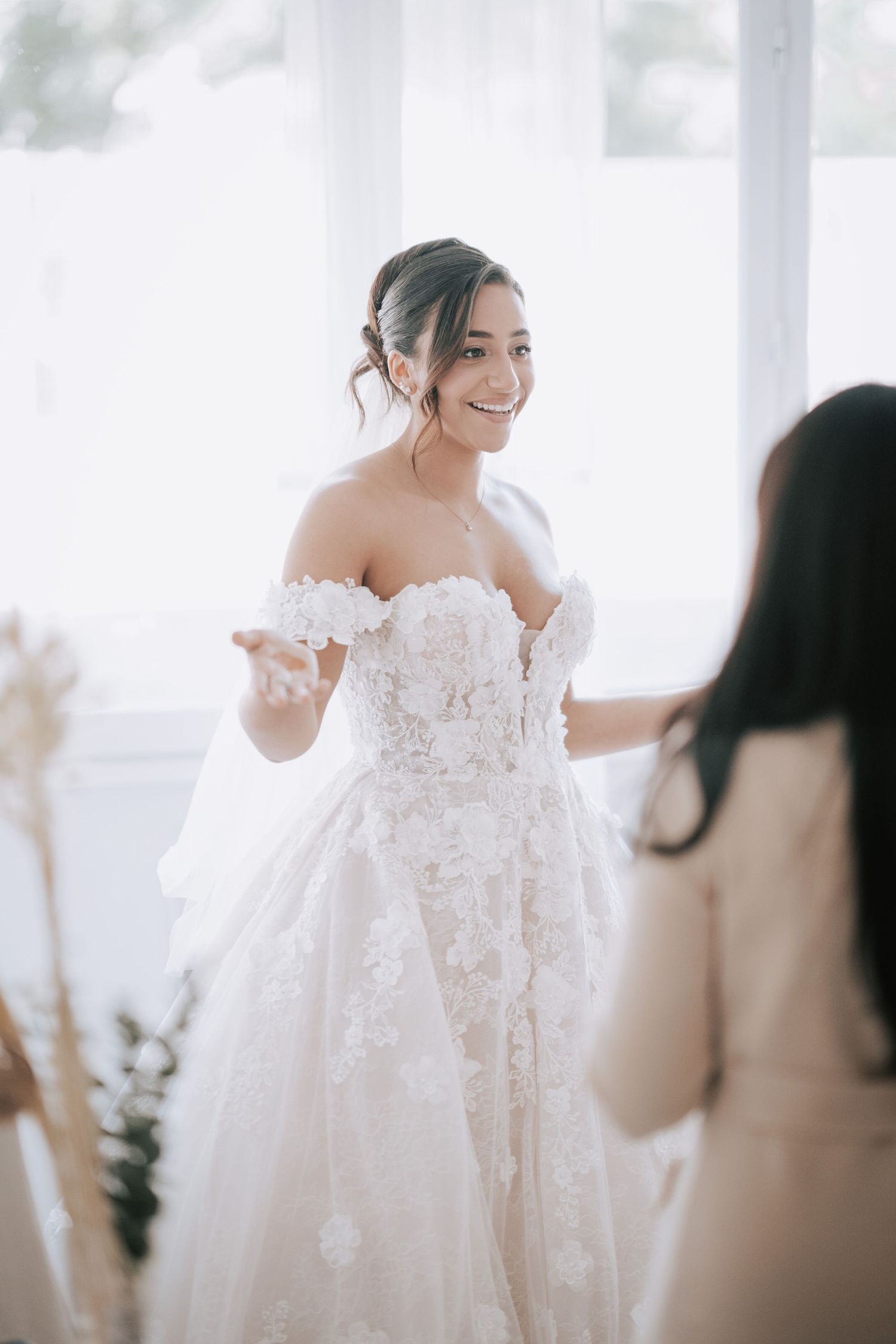 Radiant bride in off-shoulder floral lace ballgown smiles while being helped by bridesmaid in bright room.