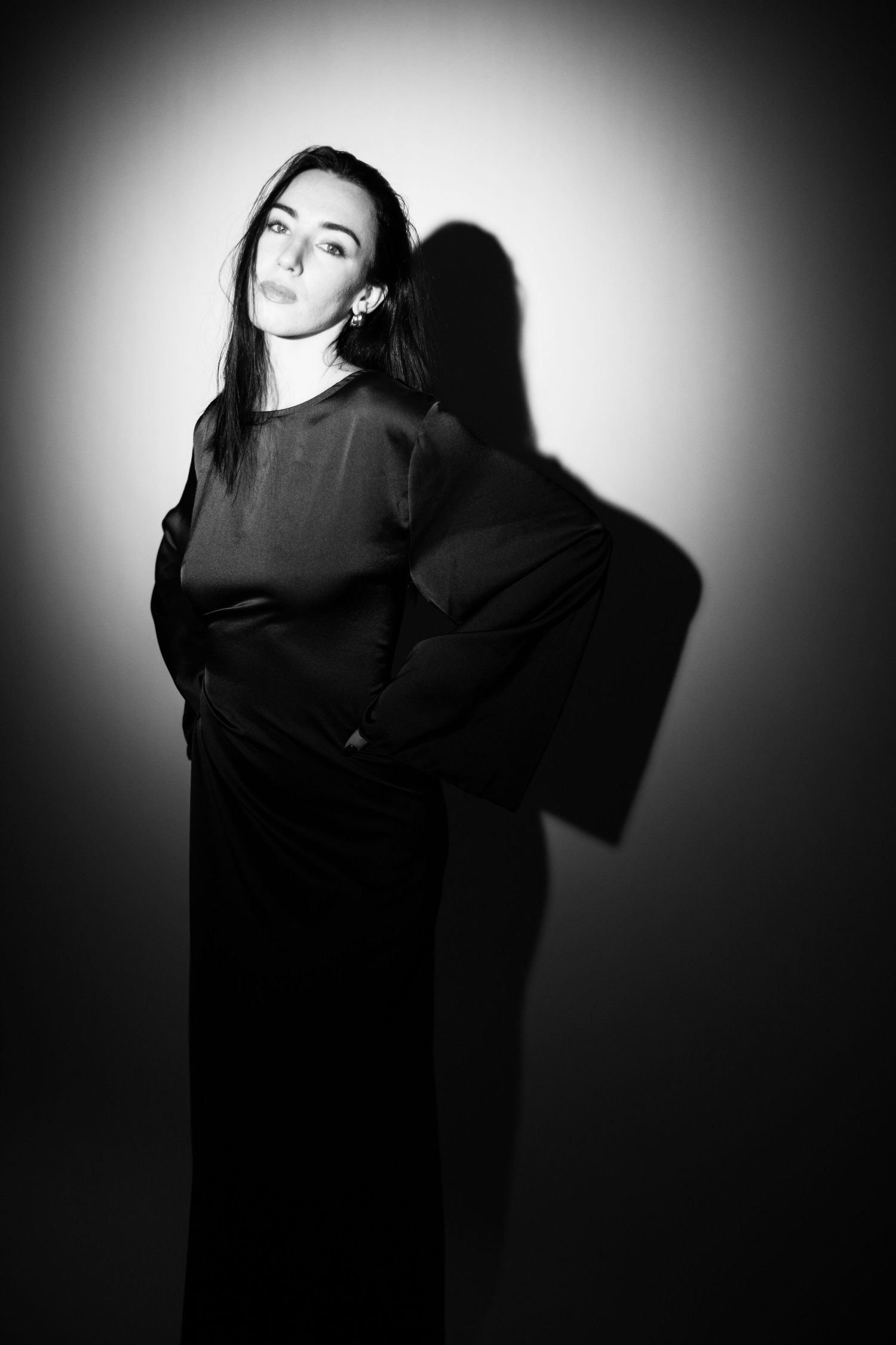 Black and white portrait of a young woman in a black outfit standing against a spotlight wall with dramatic shadows.