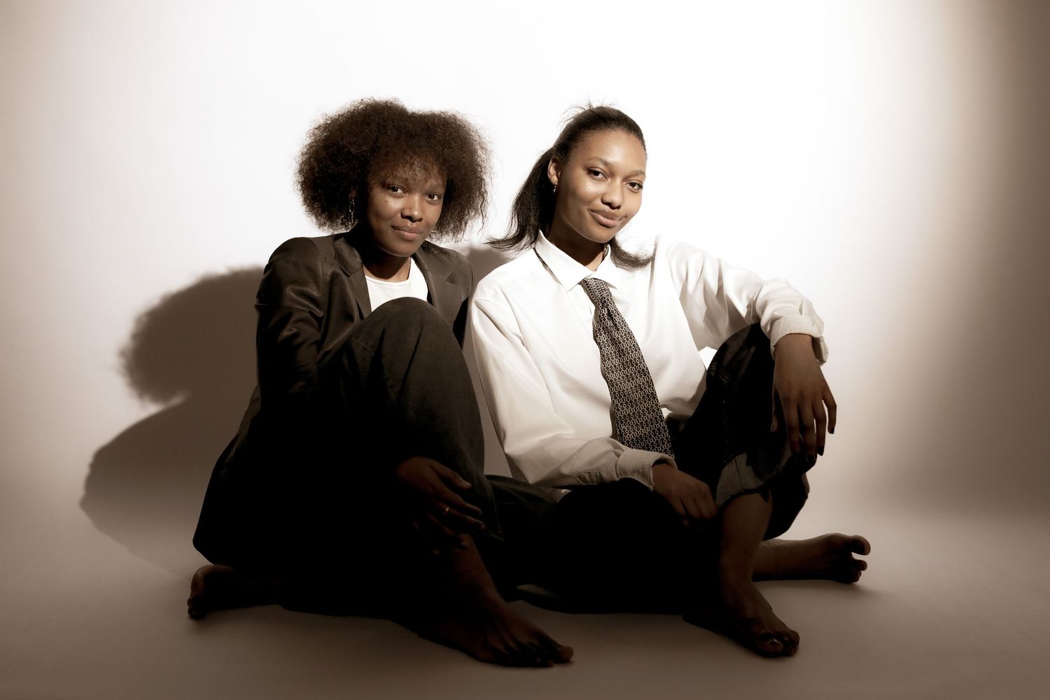 Two young women in formal attire sit together on a white floor in a stylish sepia-toned portrait photo.
