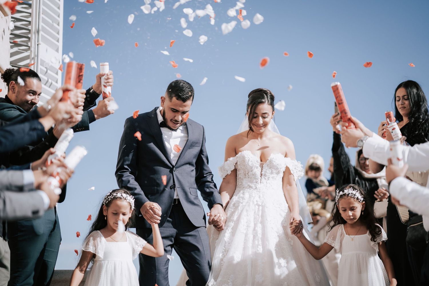 Newlyweds walk through confetti shower with flower girls on sunny wedding day celebration.