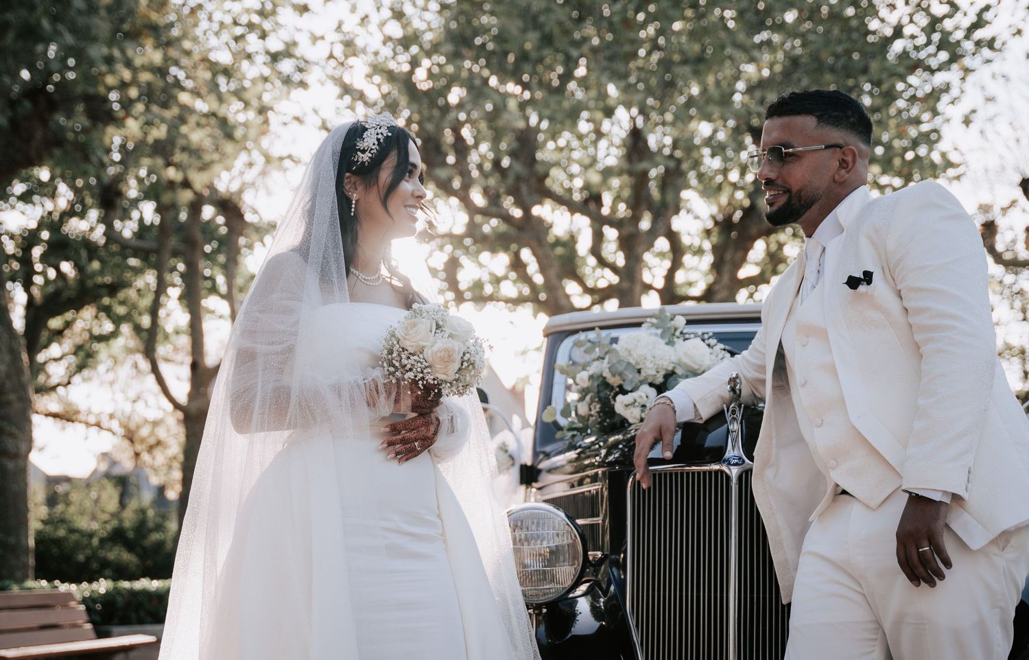 Bride in white gown and groom in ivory suit stand beside a vintage black car outdoors.