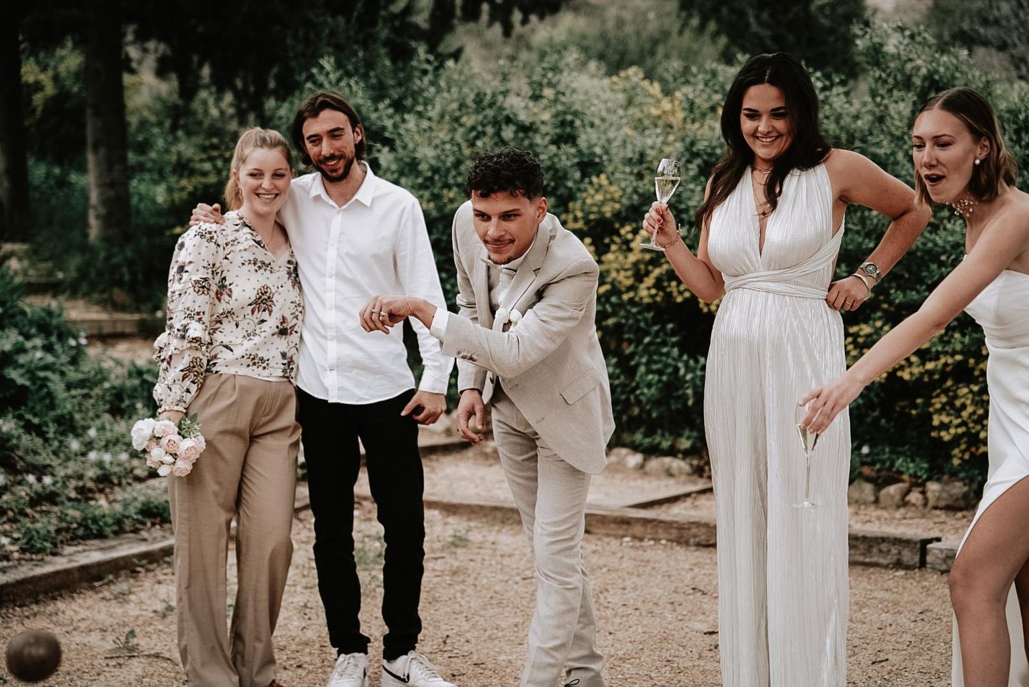 Wedding guests laugh and play petanque together in a sunny garden, mixing casual and formal attire.