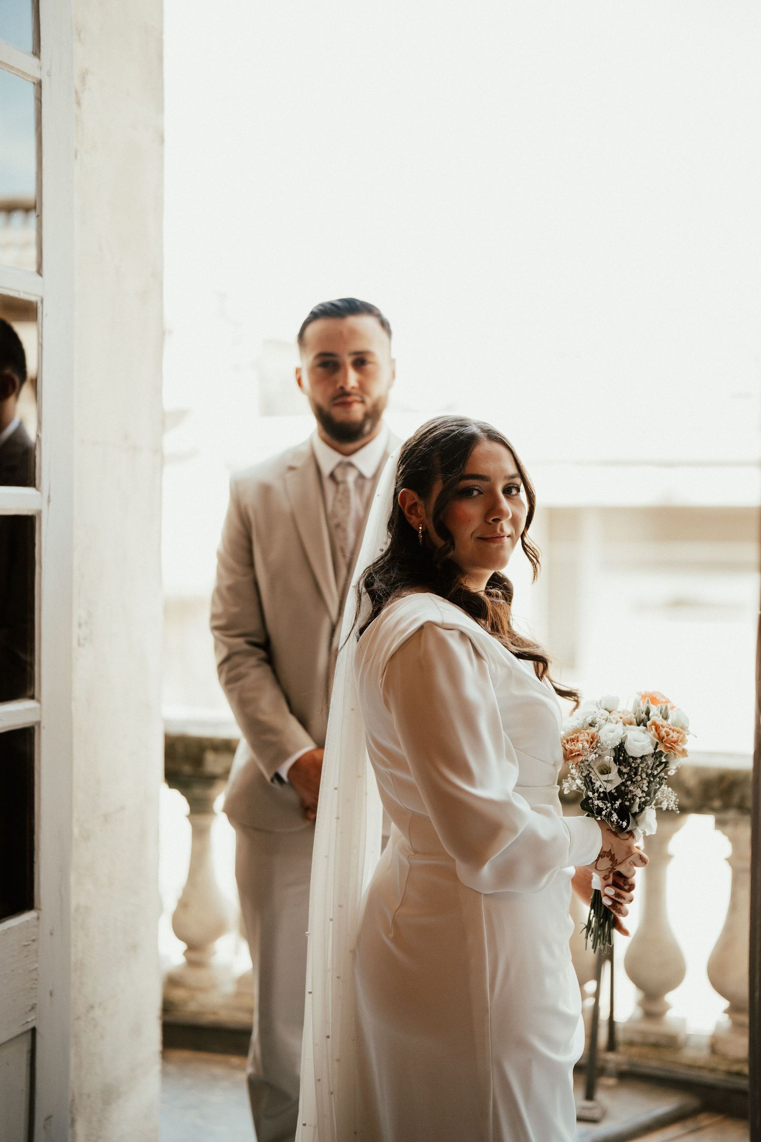 Bride smiling with bouquet while groom stands behind her on a sunlit balcony during wedding day.