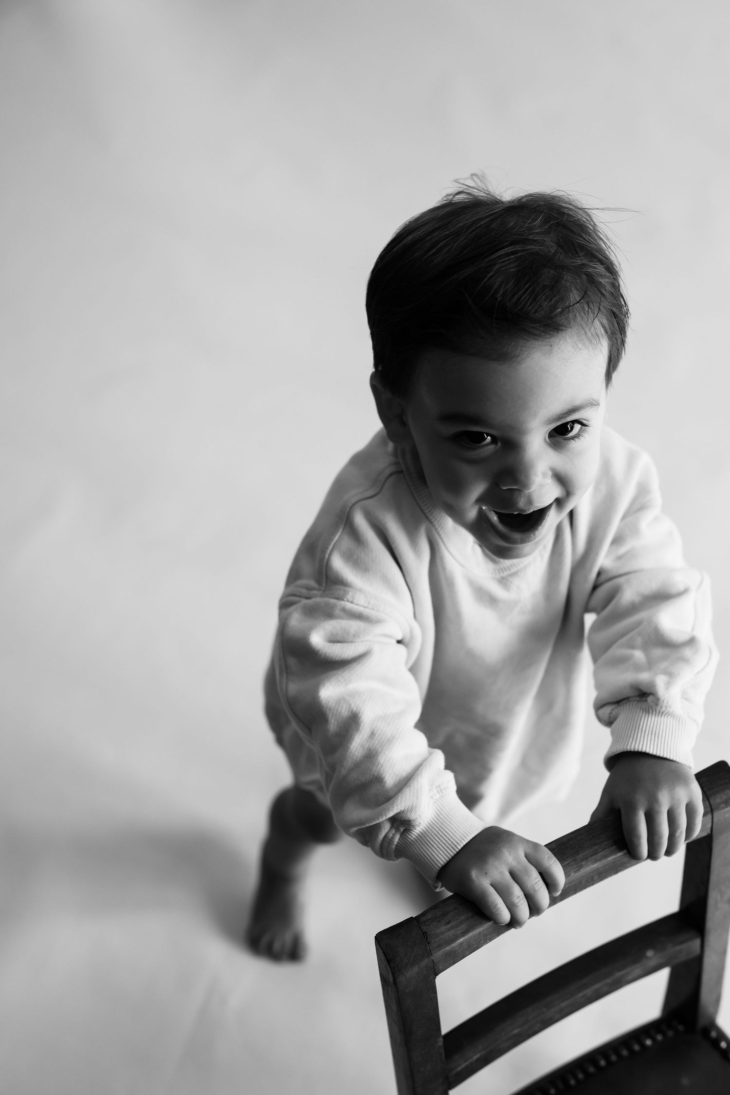 Happy laughing toddler in white sweatshirt standing by wooden chair in black and white photo.
