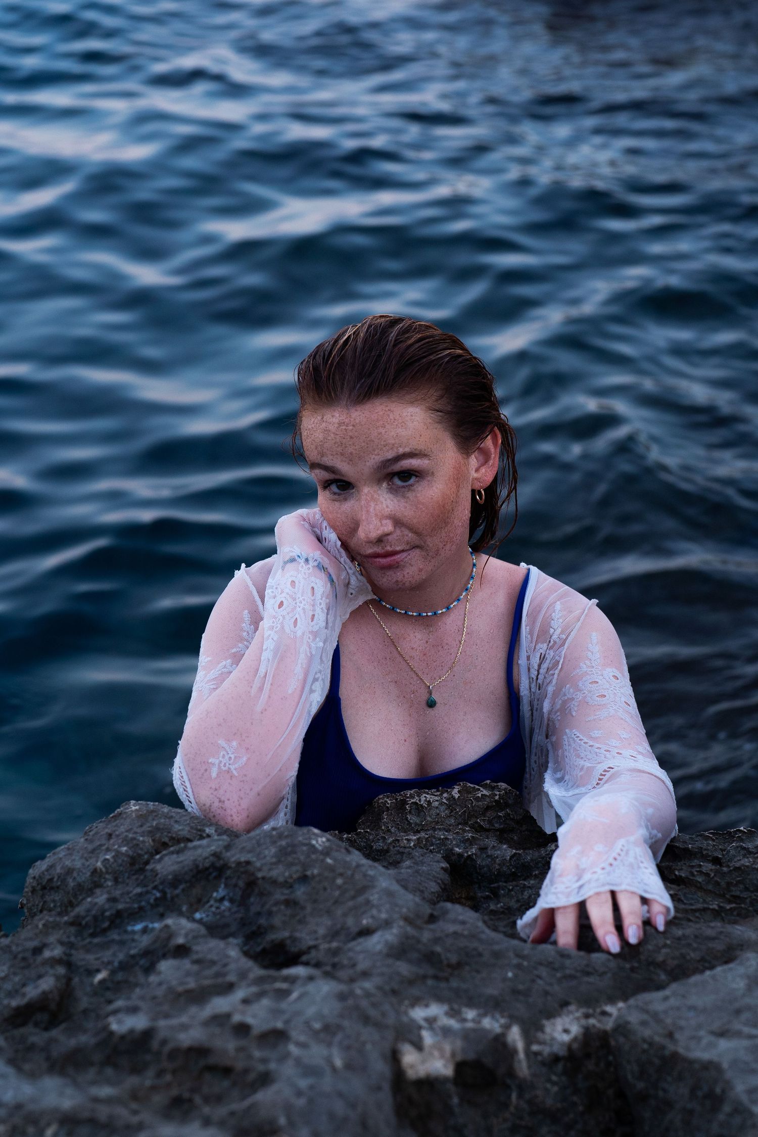 Young woman in blue swimsuit and white lace top resting on rocks in ocean water.