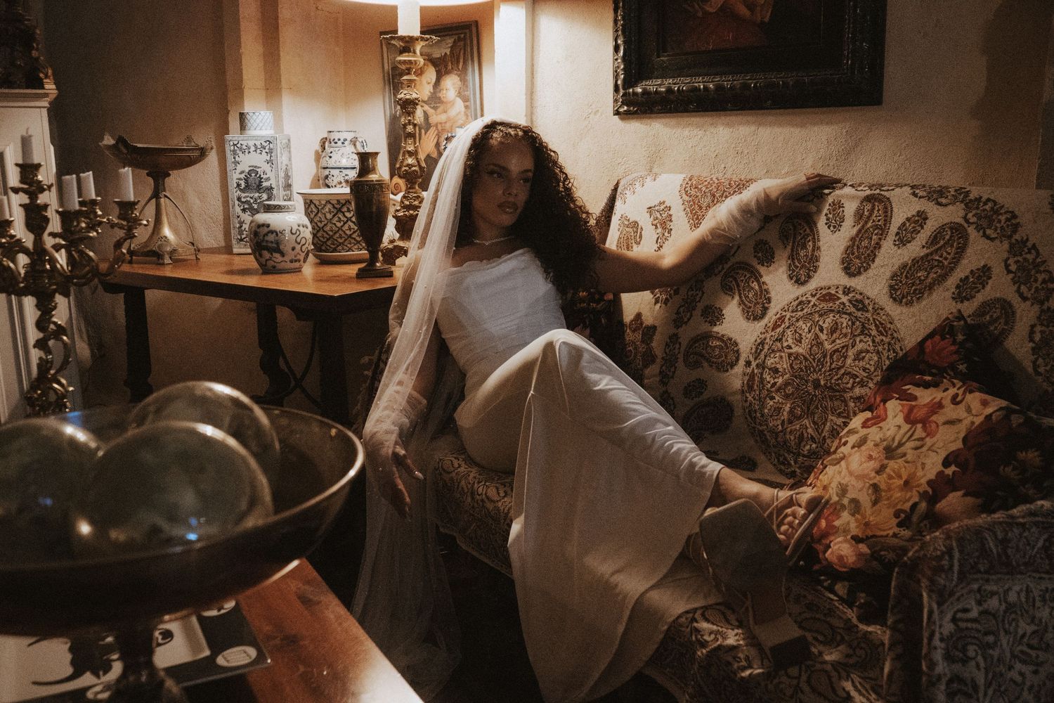 Bride in a white gown and veil reclines in an ornate vintage armchair surrounded by antique decor.