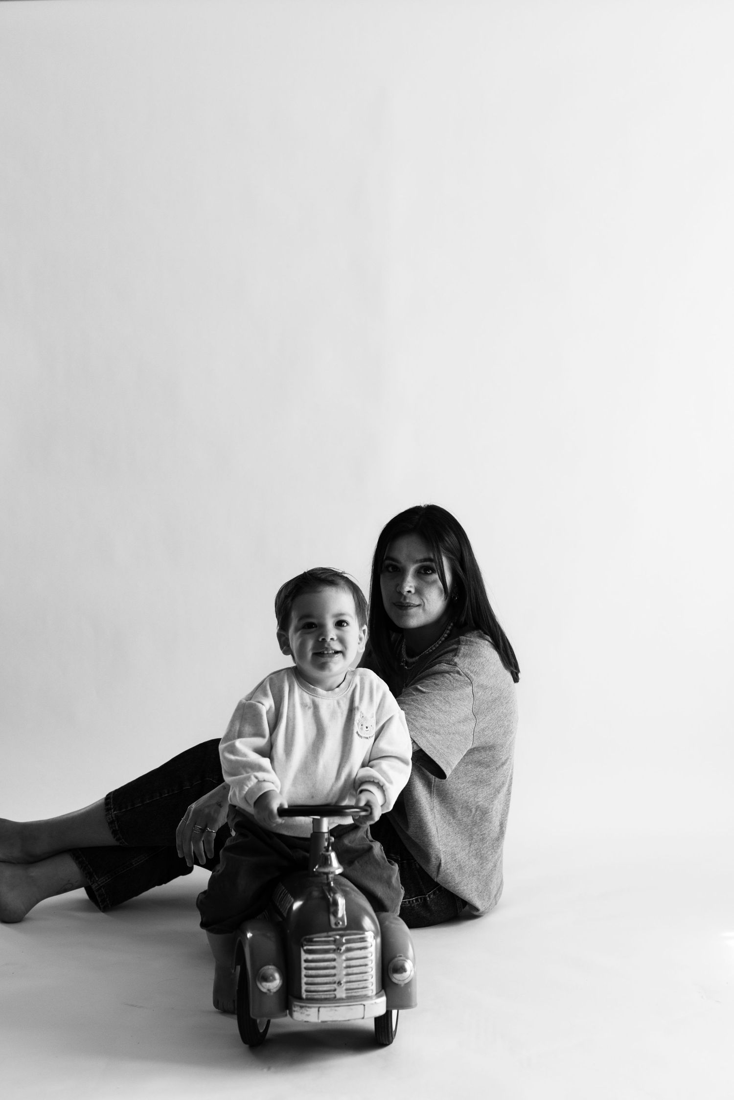 Black and white photo of a mother sitting on the floor with her smiling toddler on a toy car.