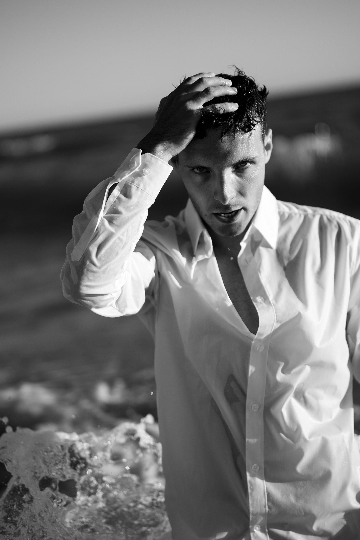 Man in wet open white shirt touching his hair while standing in ocean surf, black and white.