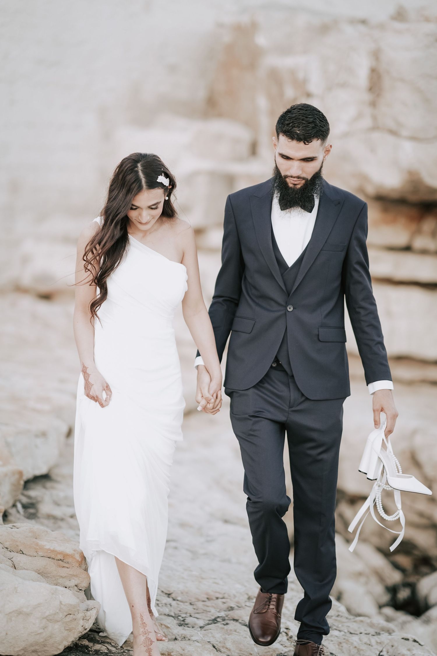 Bride and bearded groom in navy suit walk hand-in-hand on rocky terrain, groom carrying white heels.