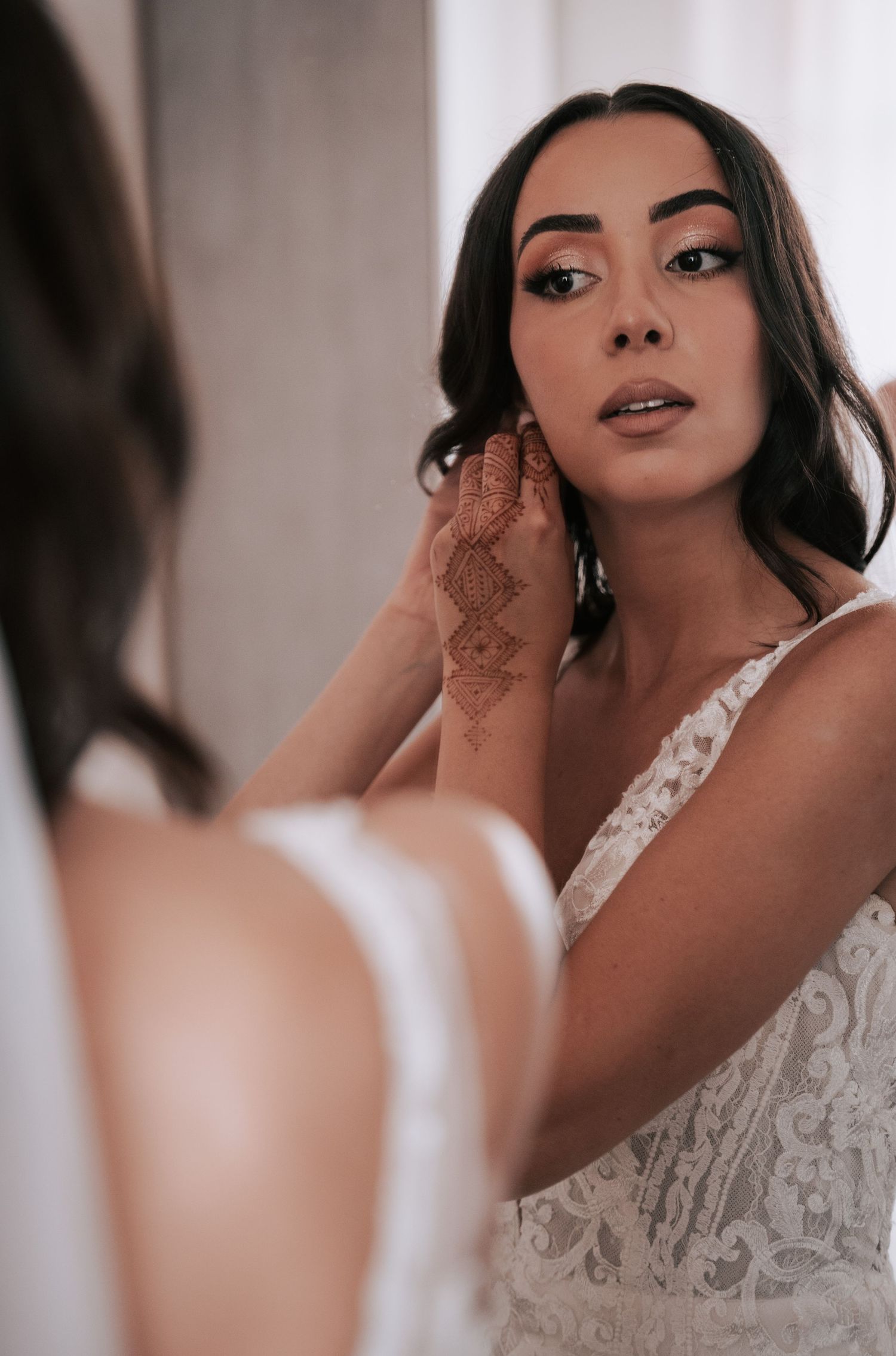 Bride with henna tattoo on hand adjusting earring while looking in mirror in lace gown.