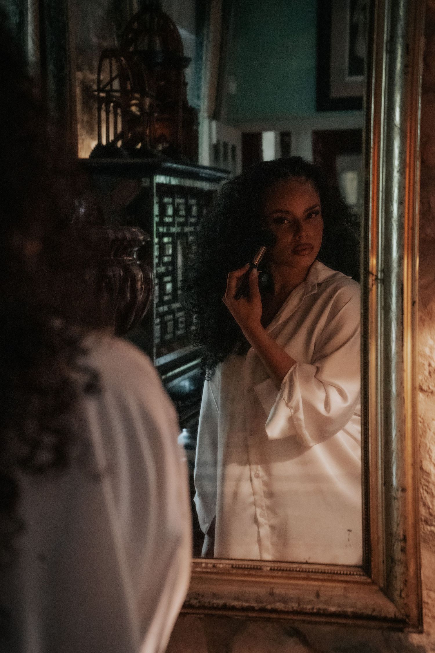 Bride with curly hair applies lipstick while gazing into an ornate vintage mirror in a decorative room.