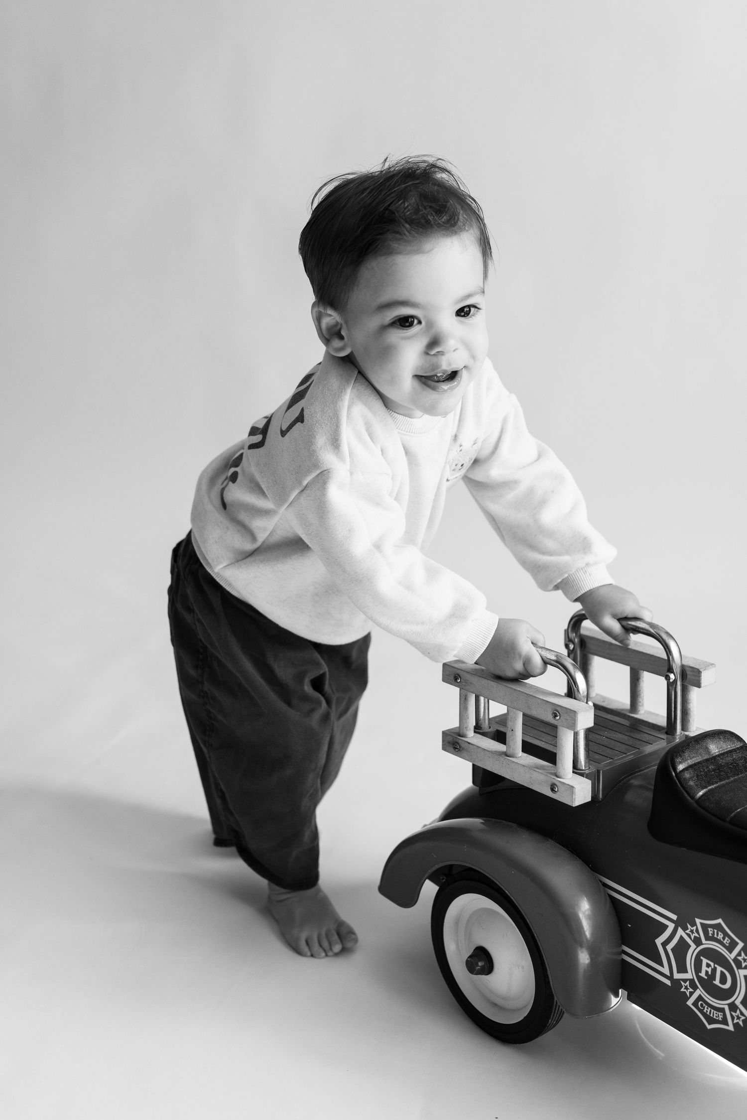 Black and white photo of a smiling toddler pushing a vintage toy fire truck on a studio floor.