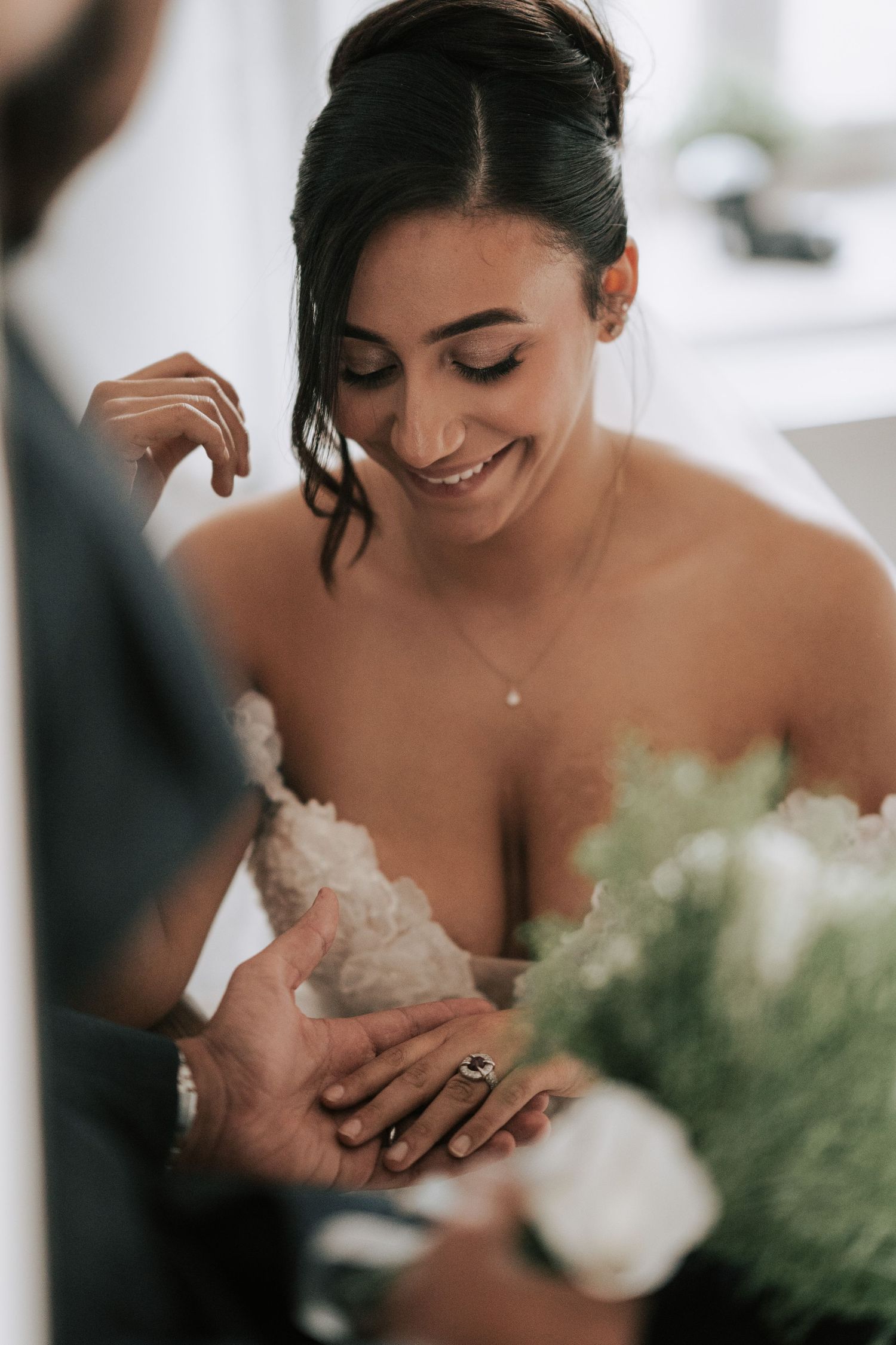Radiant bride in lace gown smiles as groom holds her hand with white floral bouquet nearby.