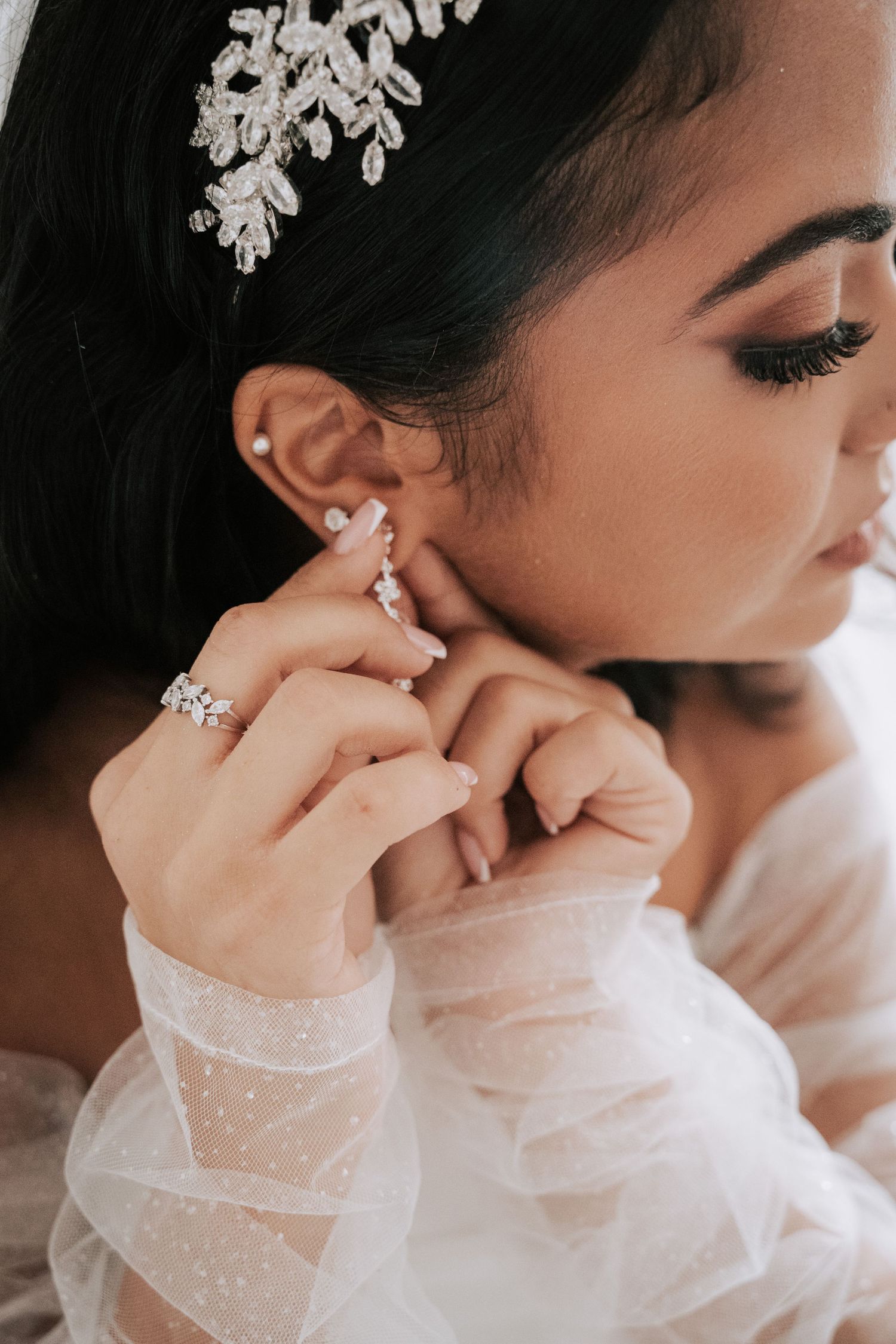 Bride putting on diamond earring with crystal headpiece and engagement ring on wedding day.