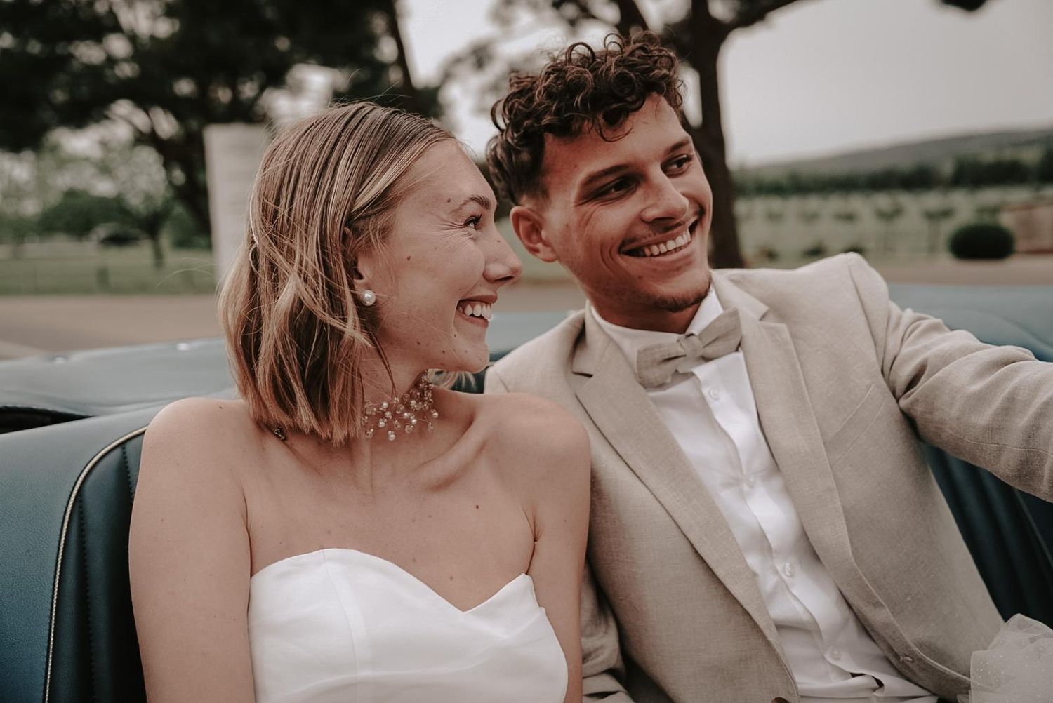 Smiling bride and groom sitting together in a vintage convertible car on their wedding day.