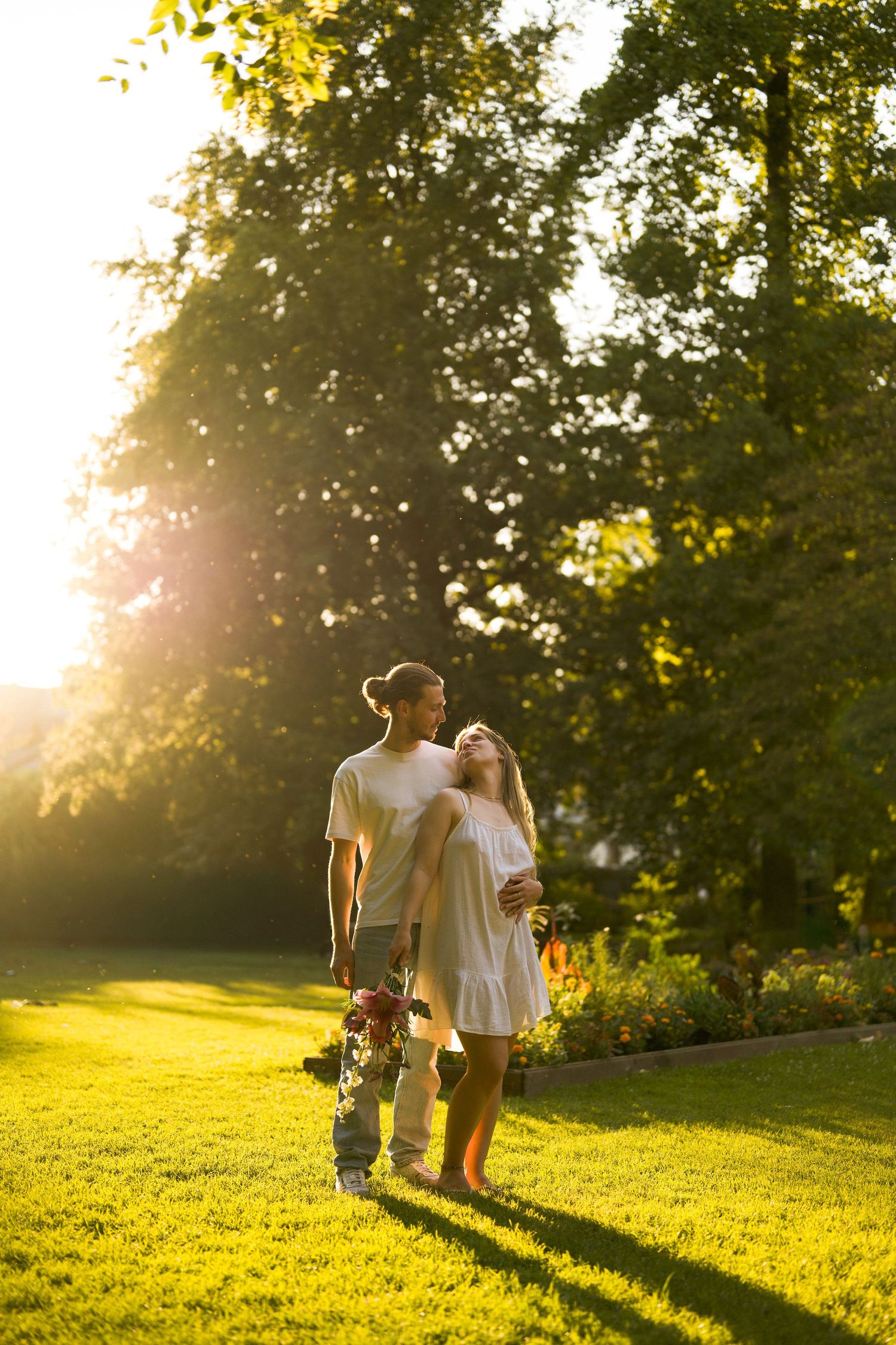 Happy couple walking hand in hand on green grass in a sunlit park holding a bouquet of flowers.