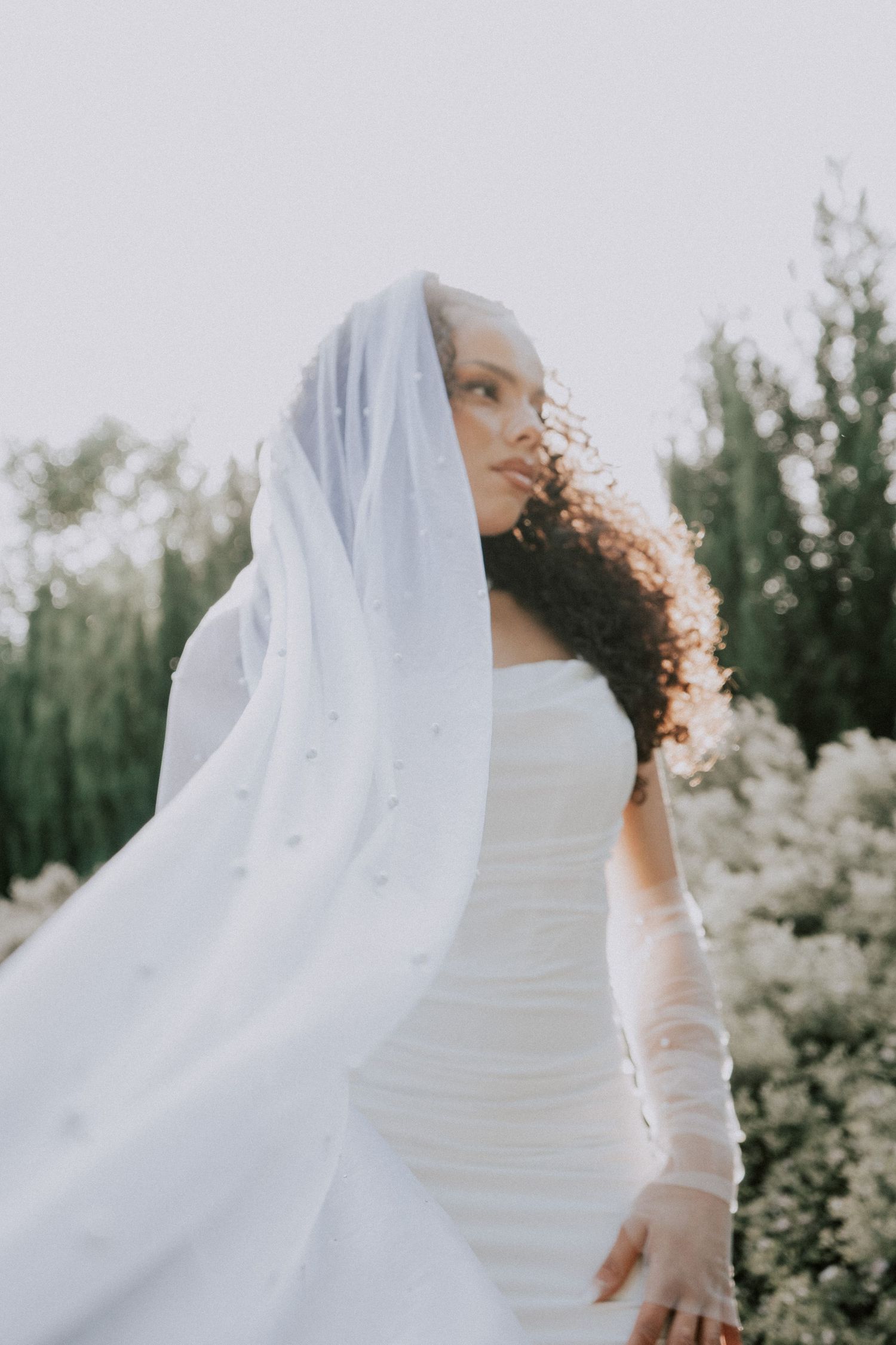 Bride in white ruched gown and flowing veil with pearl details, curly hair, posing outdoors among lush greenery.