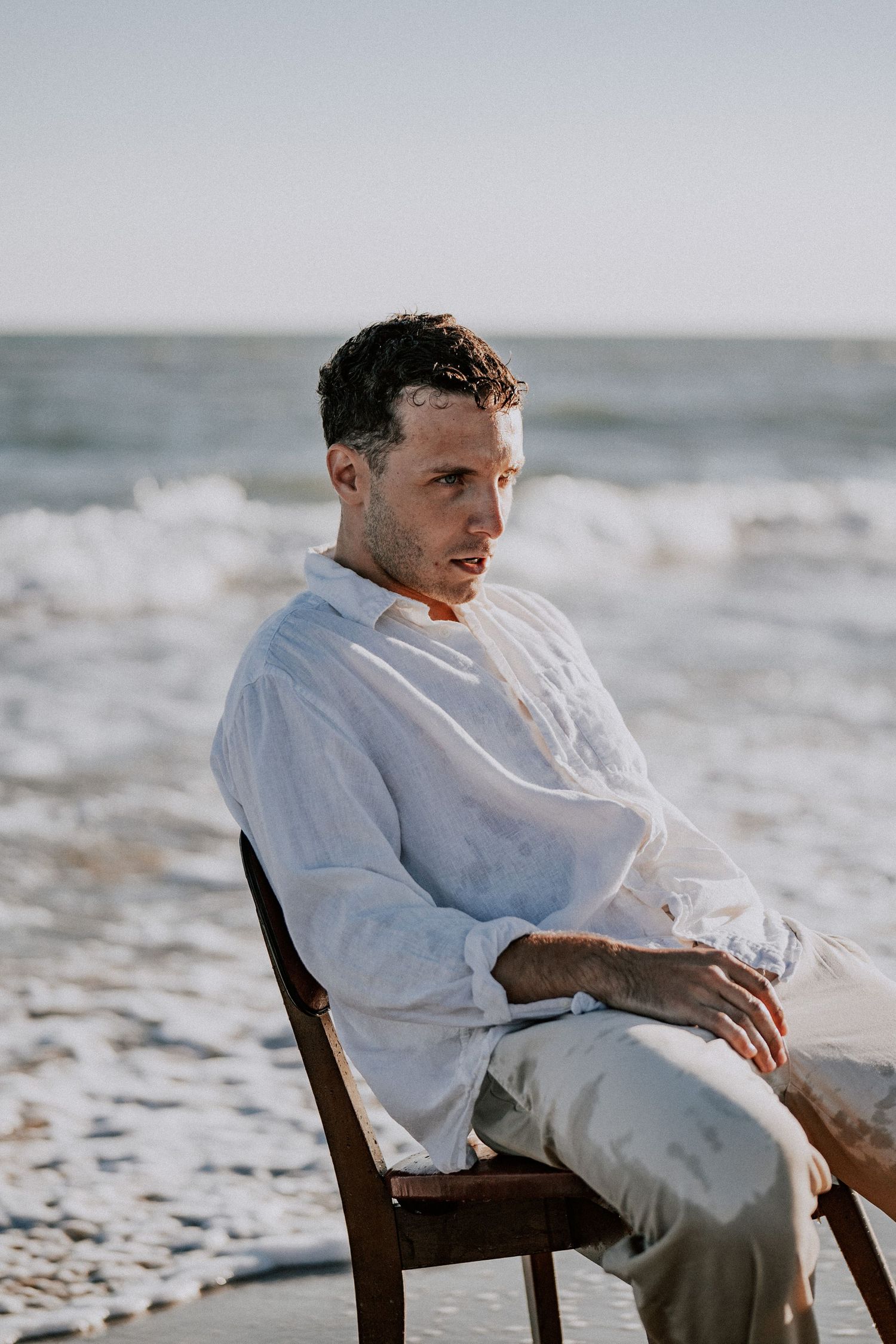 Man in wet white linen shirt sitting on wooden chair on sunny beach shoreline.