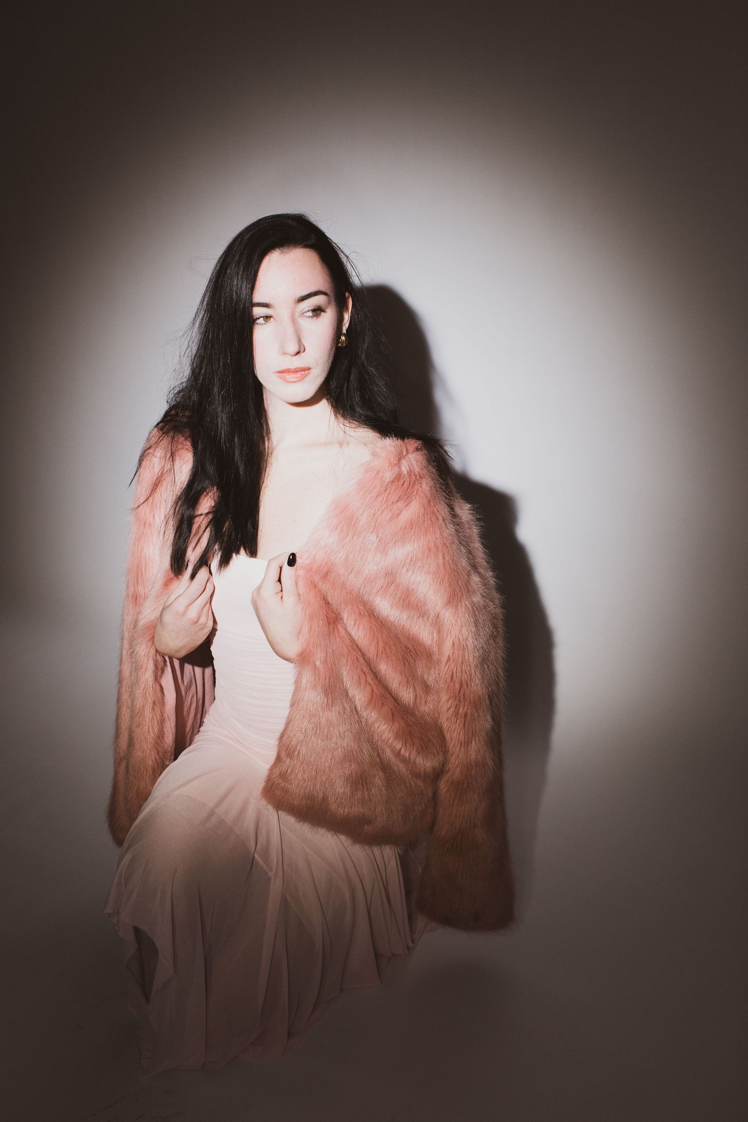 A young woman with dark hair poses in a pink fur coat and light dress against a softly lit white background.