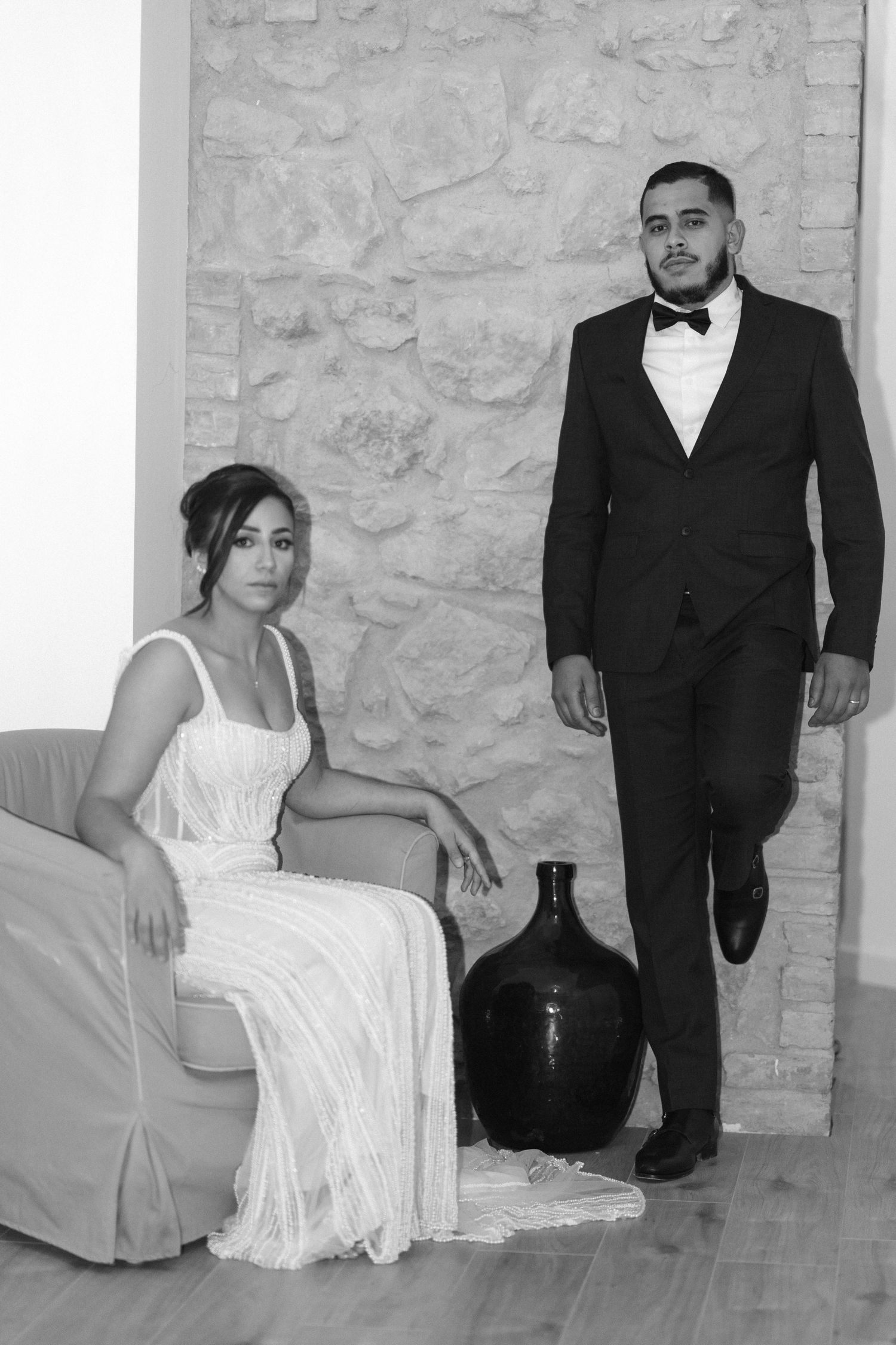 Black and white formal wedding portrait of bride seated in chair and groom standing beside a stone wall backdrop.