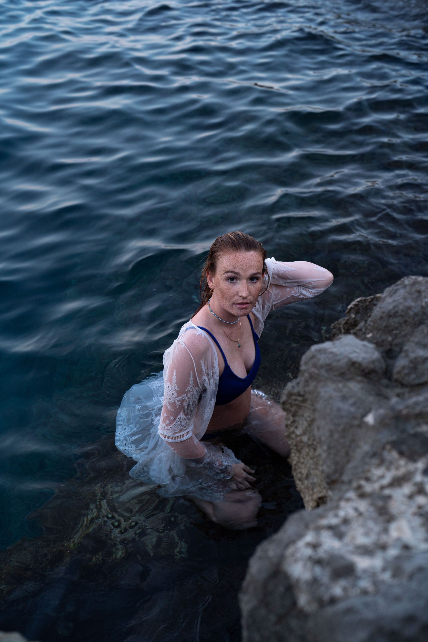 Woman in blue bikini and white lace cover-up sitting on rocks in the ocean at dusk.