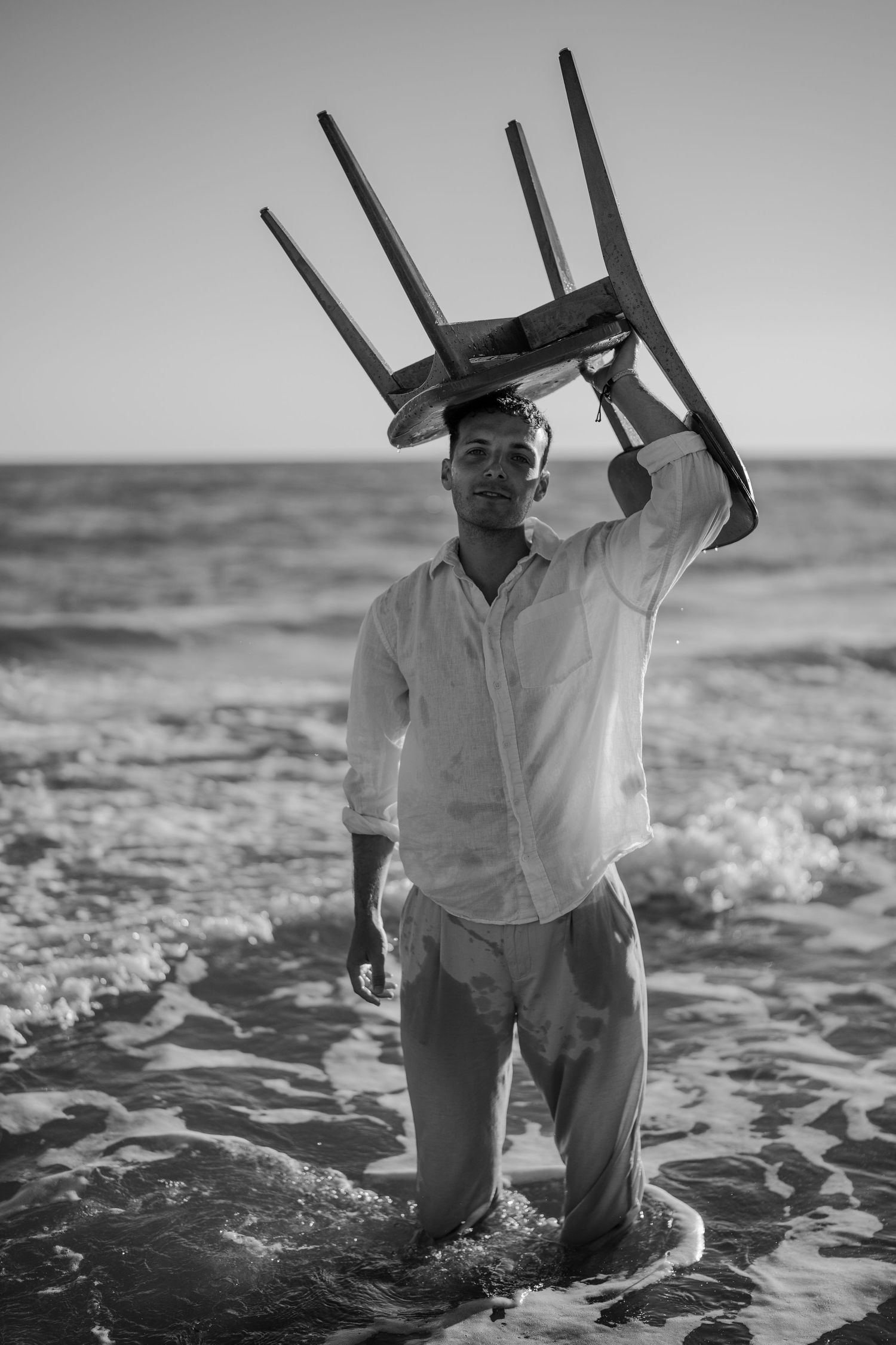Man in white shirt carrying wooden chair overhead while standing in ocean waves, black and white.