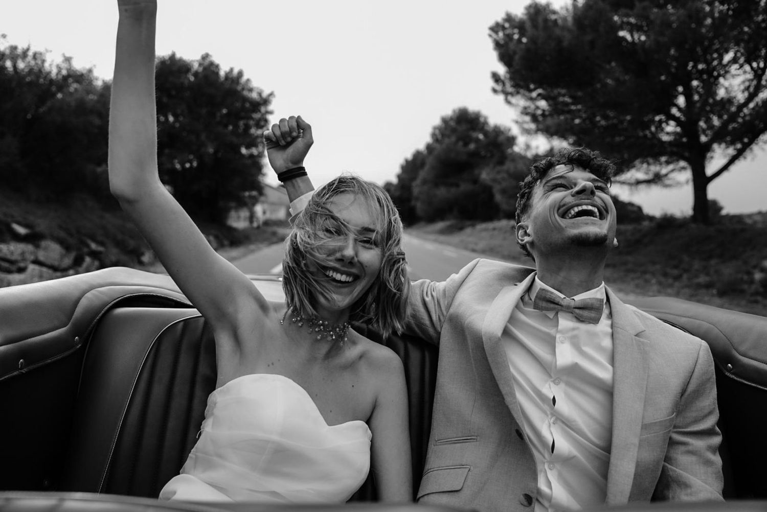 Joyful bride and groom celebrating in a vintage convertible car, black and white wedding photo.