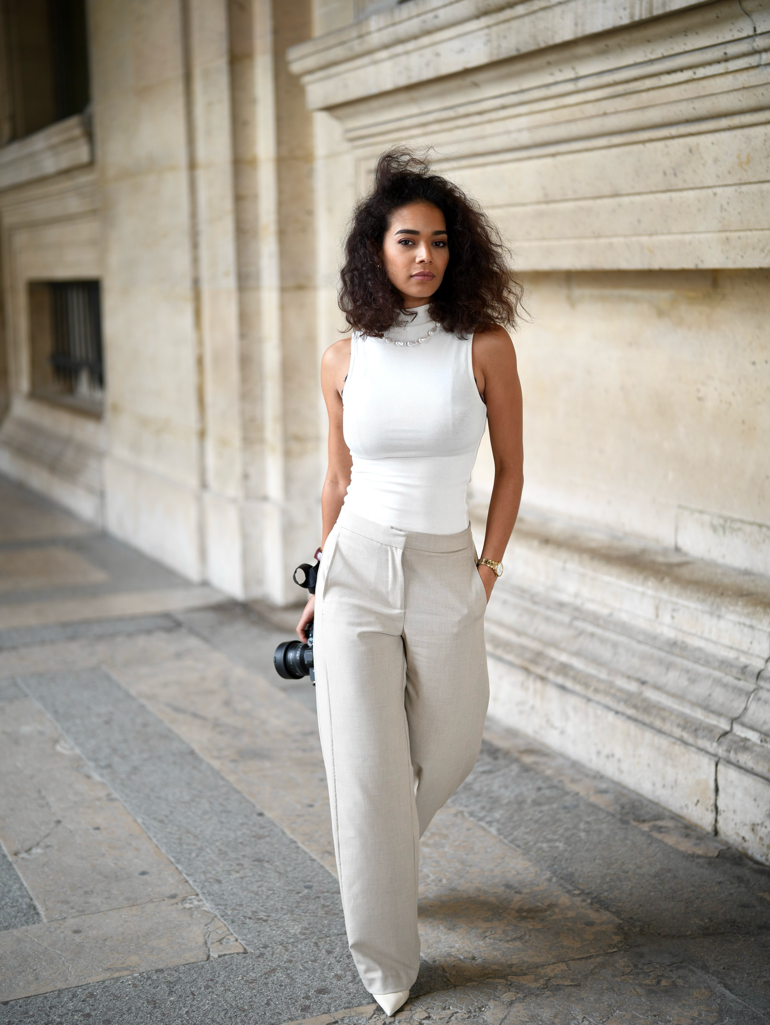 A young woman poses outdoors in a white sleeveless turtleneck top and wide-leg cream trousers near a stone building.